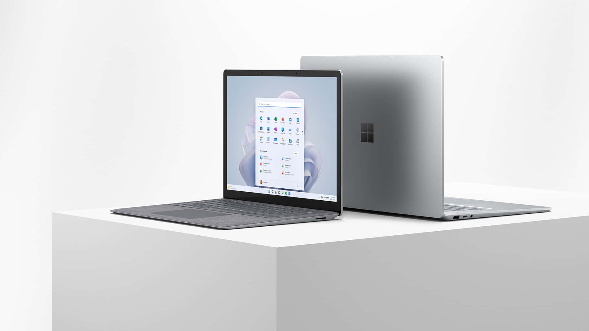 為您便利而設計的流線型筆記本電腦 壁紙 — 兩部時尚的銀色 Microsoft Surface 筆記型電腦展示在白色底座上，一部打開顯示開始功能表，另一部關閉，極簡工作室拍攝。