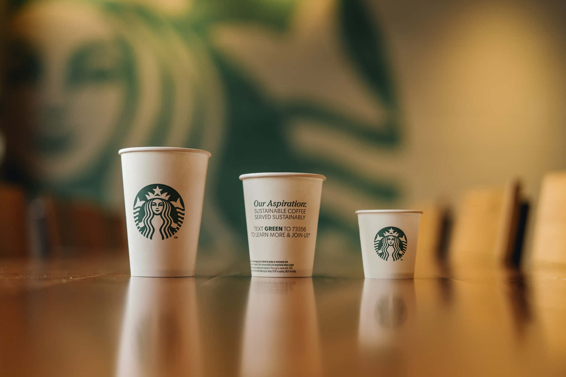 三個Starbucks咖啡杯排列在木製櫃檯上，背景是綠色標誌牆。 — 星巴克 壁紙
