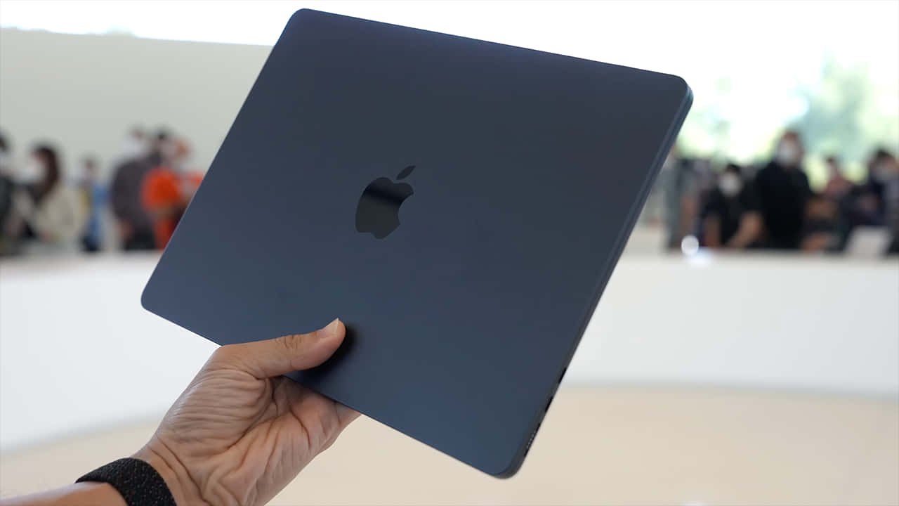 在產品活動中手持深藍色MacBook，背景是模糊的觀眾。