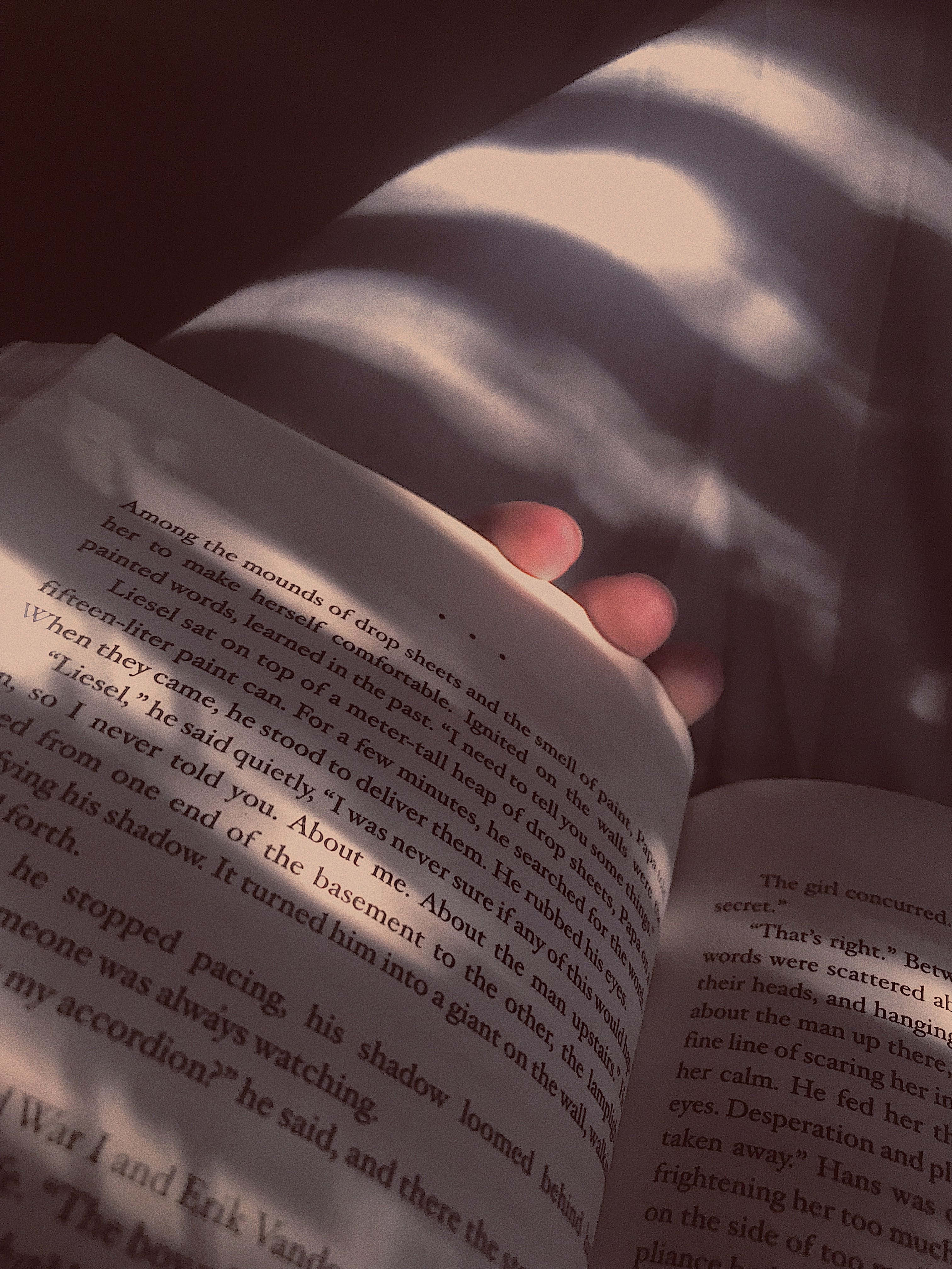為您精心排列的美學書籍陣列 壁紙 — 一隻手拿著一本打開的書，書上文字清晰可見，柔和陽光在頁面上投下陰影。