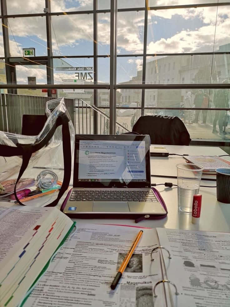 為成功而準備！ 壁紙 — 一部手提電腦放在雜亂的書桌上，有打開的筆記本和一杯水，透過大窗戶看到明亮的城市景觀。