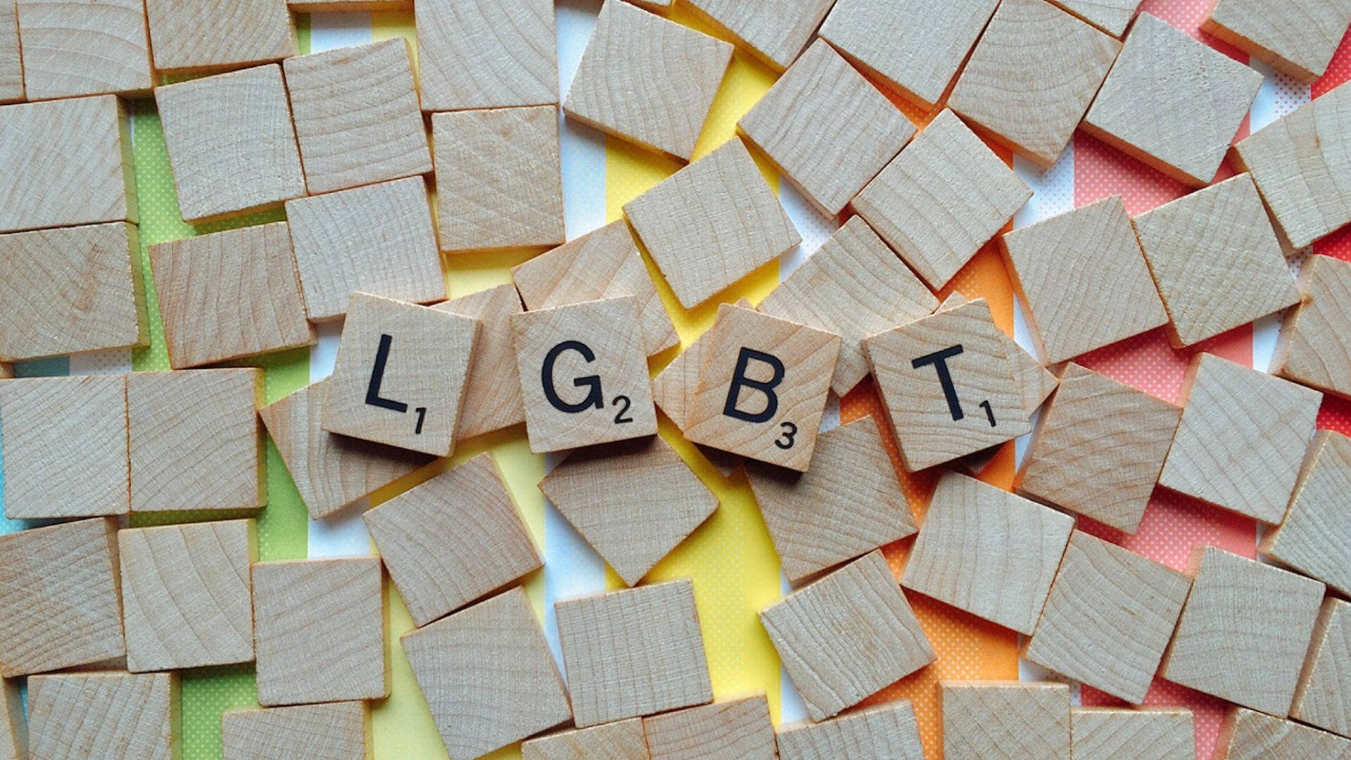 為我們的LGBT+家庭獻上的簡約彩虹 壁紙 — 木製 Scrabble 字母拼出 LGBT，排列在彩色條紋背景上。