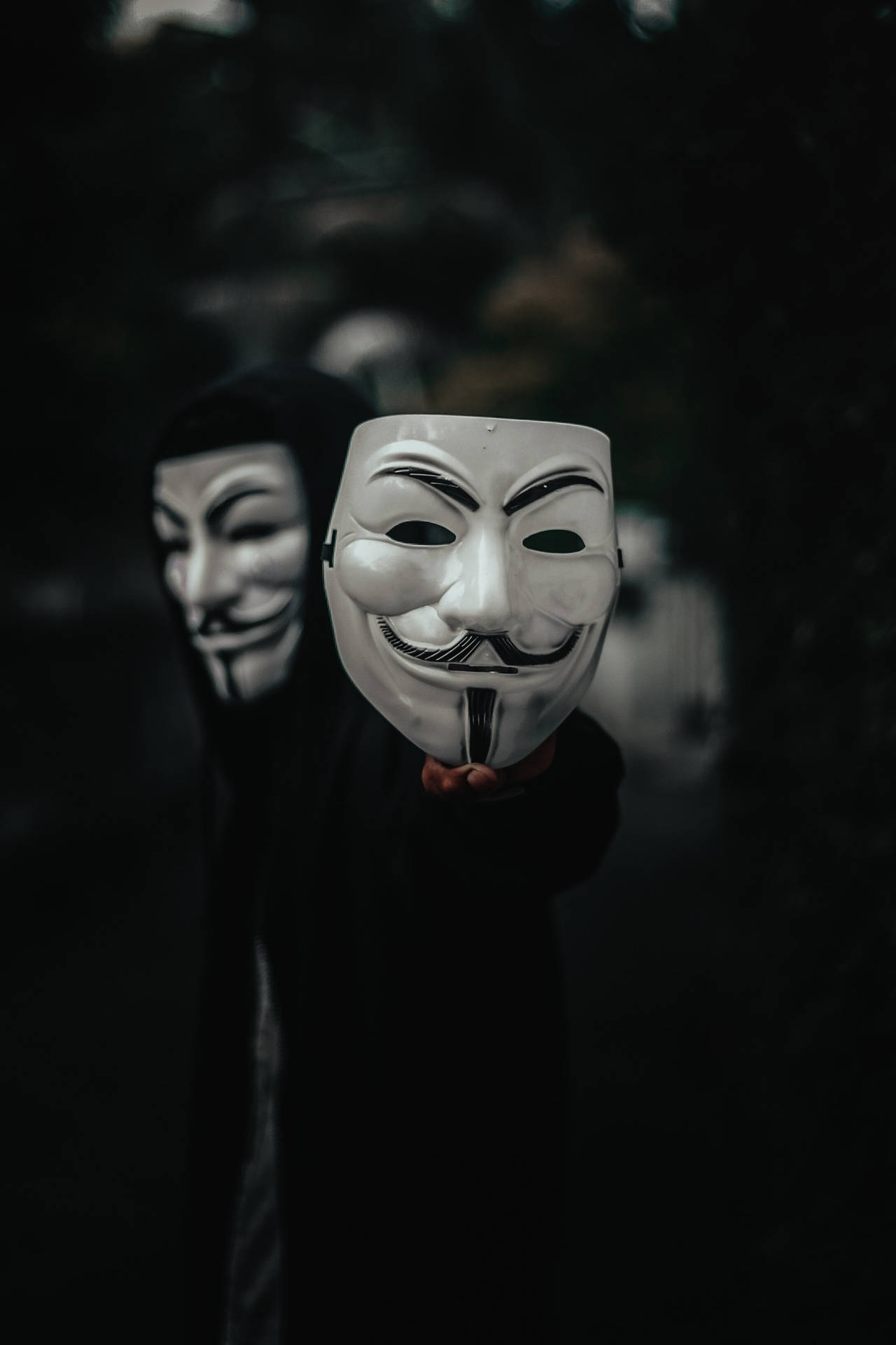穿著黑色連帽衫的人在黑暗環境中手持白色蓋伊·福克斯面具。