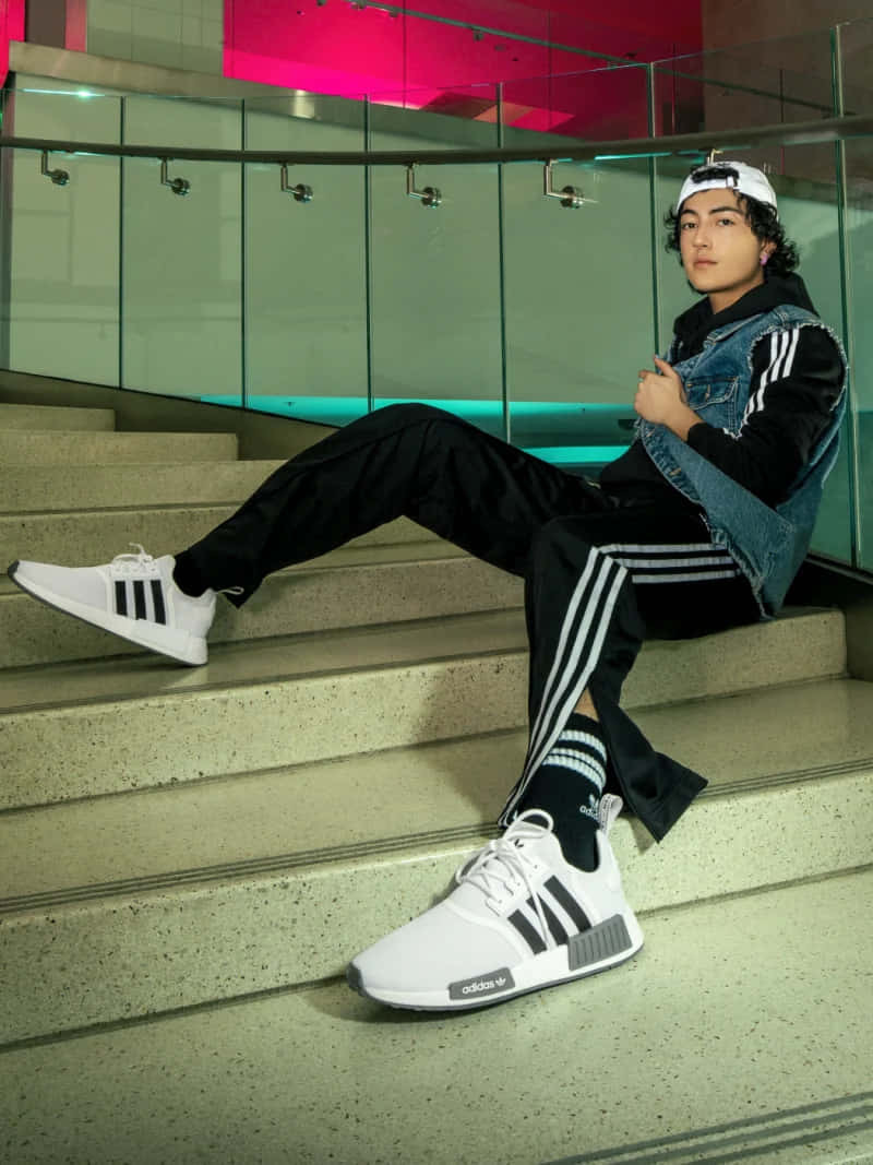為行動而生 — Adidas 壁紙 — 一名身穿白色 adidas 運動鞋和牛仔背心的年輕男子，在霓虹粉紅和藍綠色燈光下，悠閒地躺在現代混凝土樓梯上。
