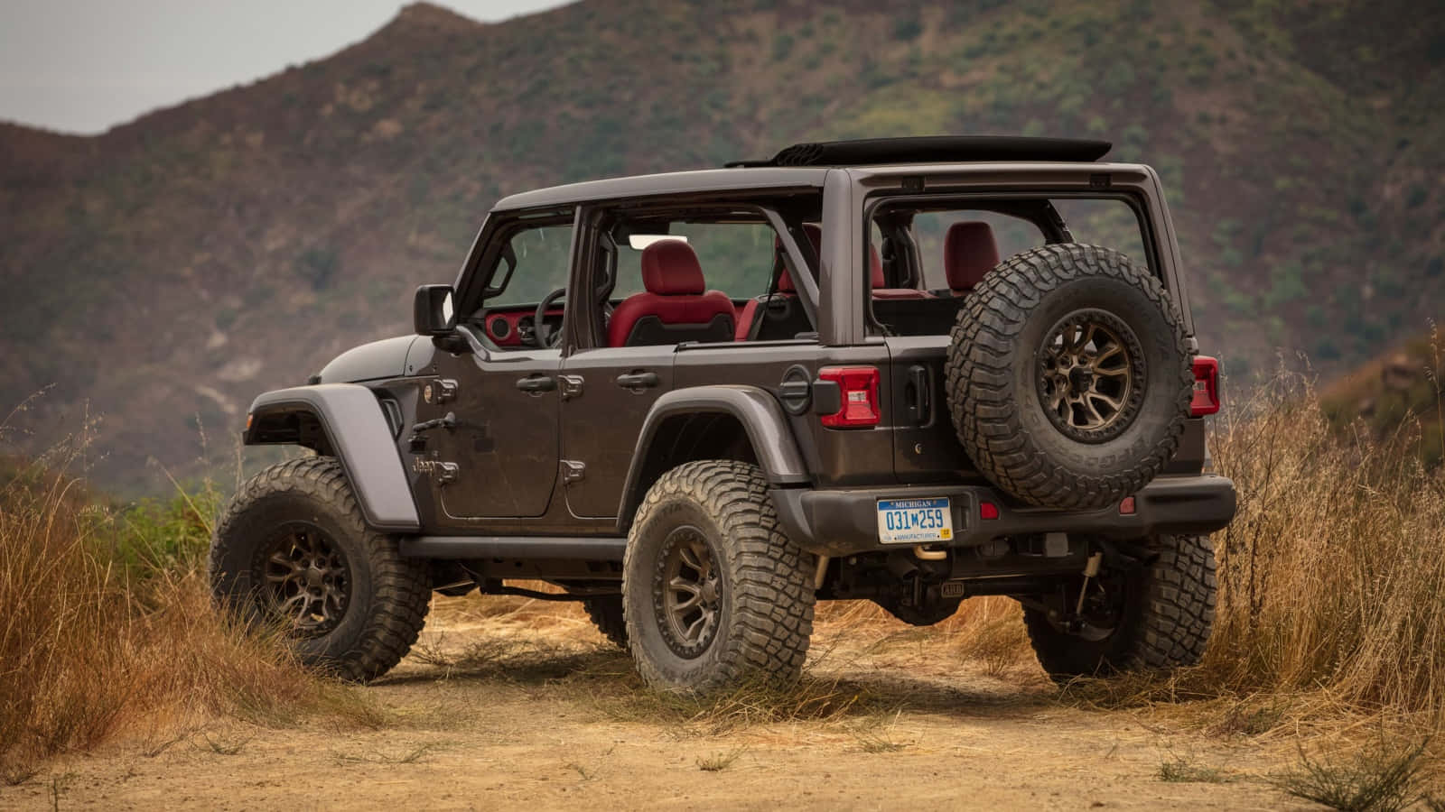 為越野愛好者準備，以經典Jeep Wrangler駛入小徑 壁紙 — 一輛堅固的深棕色Jeep Wrangler，配備紅色內飾和後置備用輪胎，在陰天下行駛於乾燥的草坡。