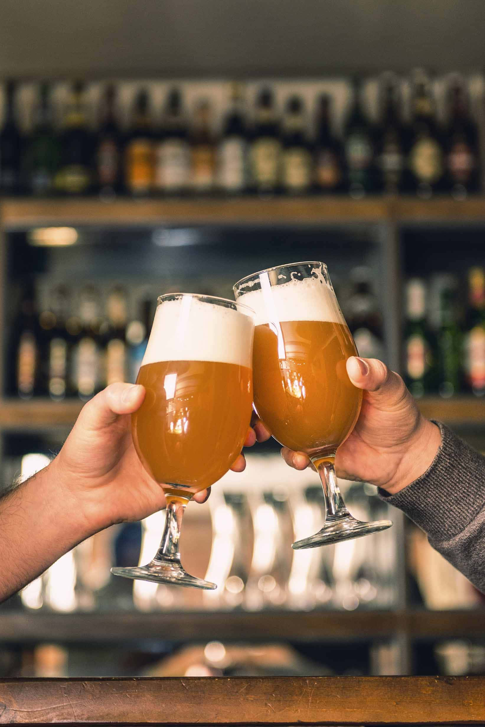 為週末乾杯！ 壁紙 — 兩隻手在溫暖燈光的酒吧中碰杯，琥珀色啤酒杯帶有泡沫，背後是模糊的酒架。