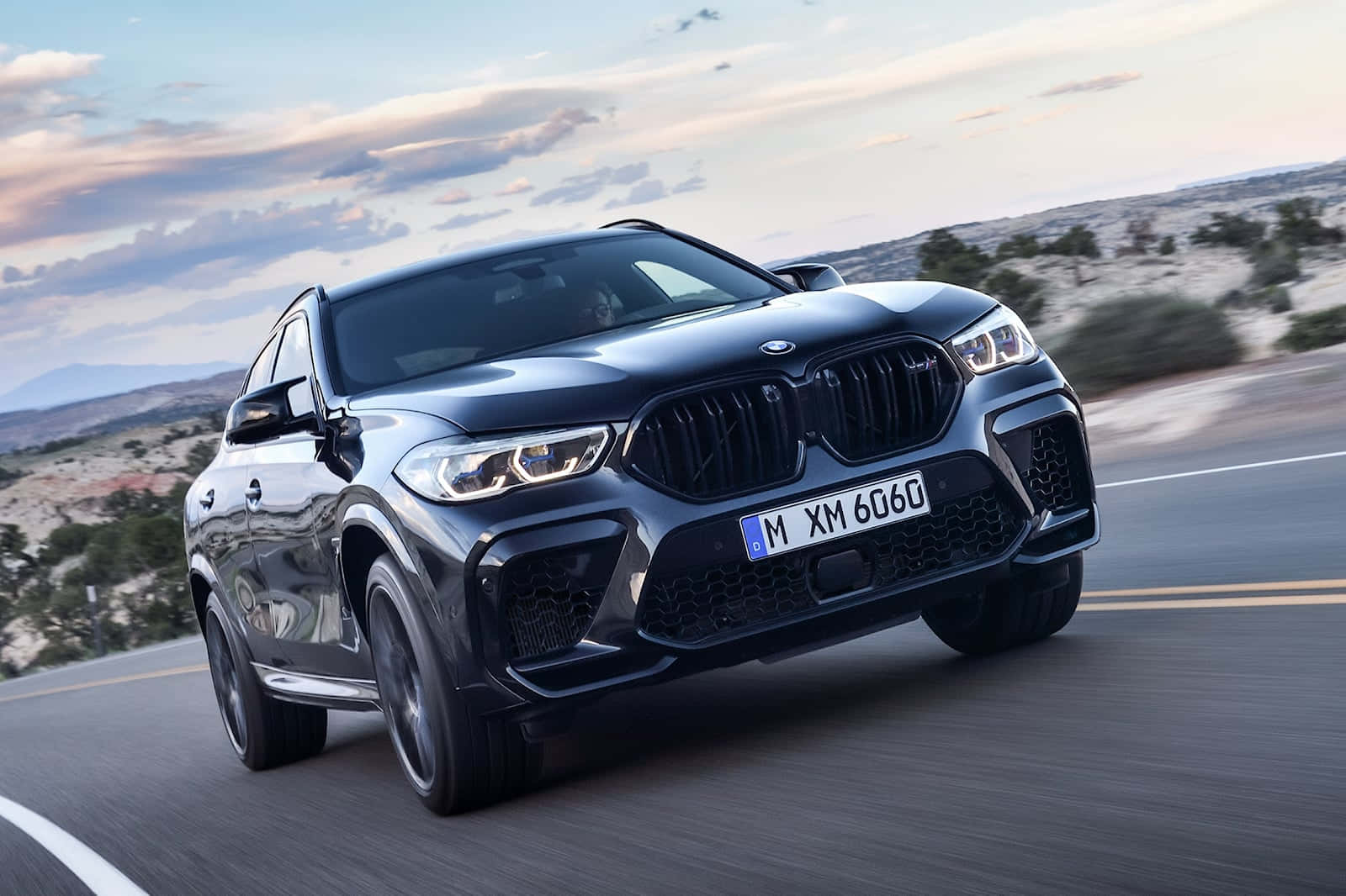 為駕馭而生 — BMW X6 M 壁紙 — 一輛時尚的黑色BMW X6M在沙漠高速公路上疾馳，天空柔和暮色，雲朵零星散落。