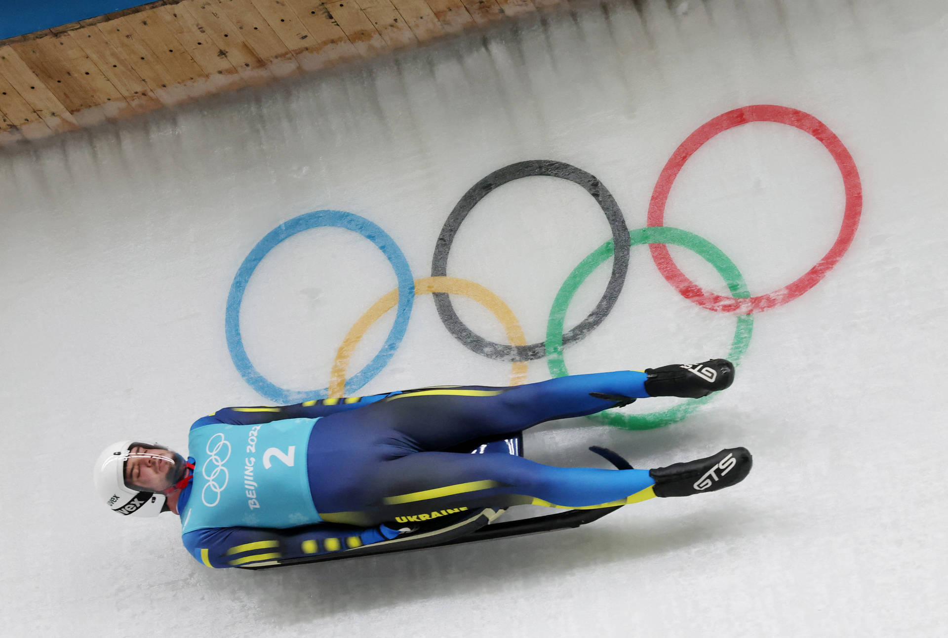 雪橇運動員在冰道上比賽，身後畫有奧運五環。