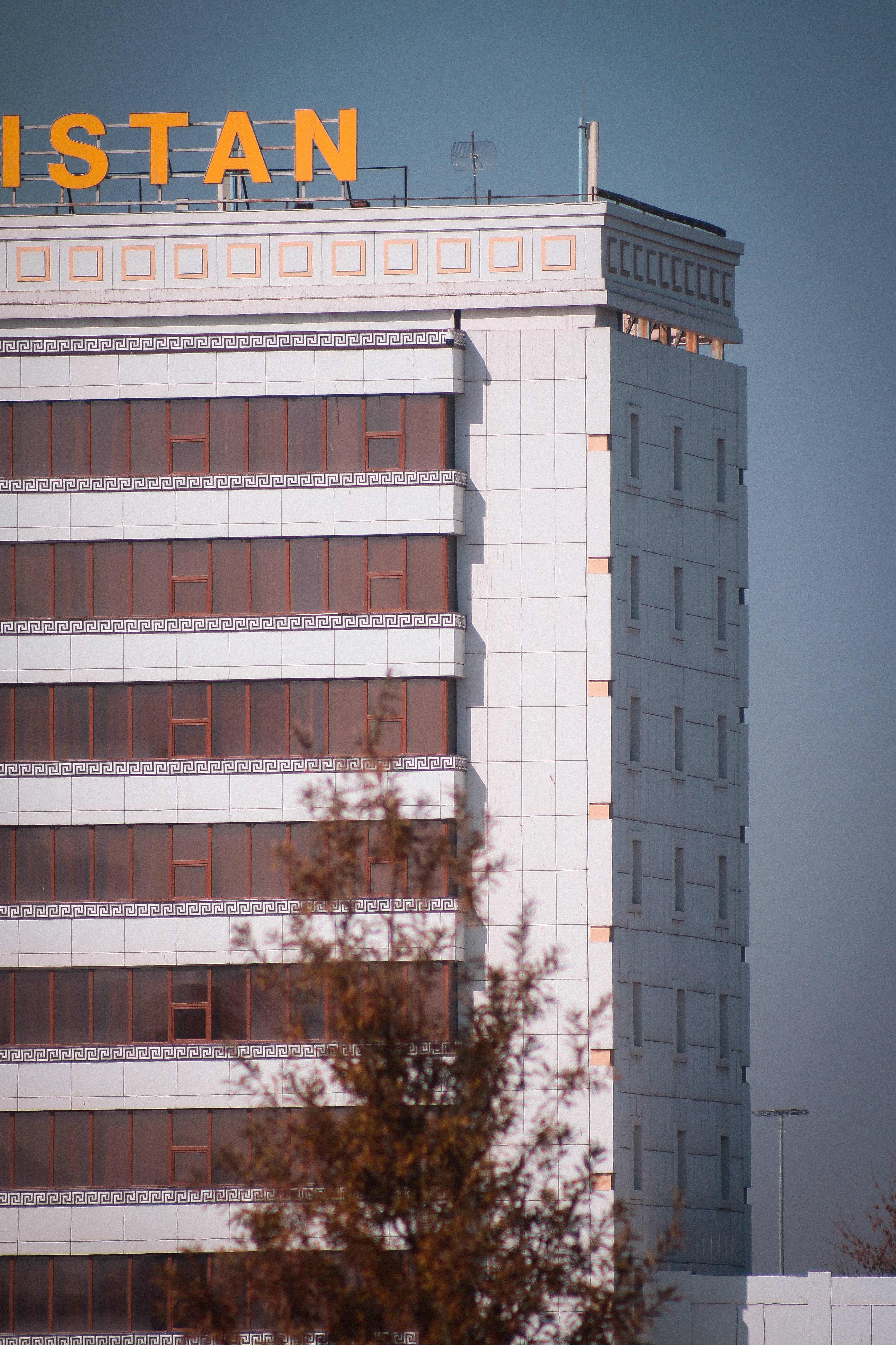 烏茲別克斯坦大飯店建築 壁紙 — 一座高聳的白色建築物，帶有橙色 'ISTAN' 標誌和幾何圖案，矗立在晴朗的藍天下，前景有秋天的樹枝。