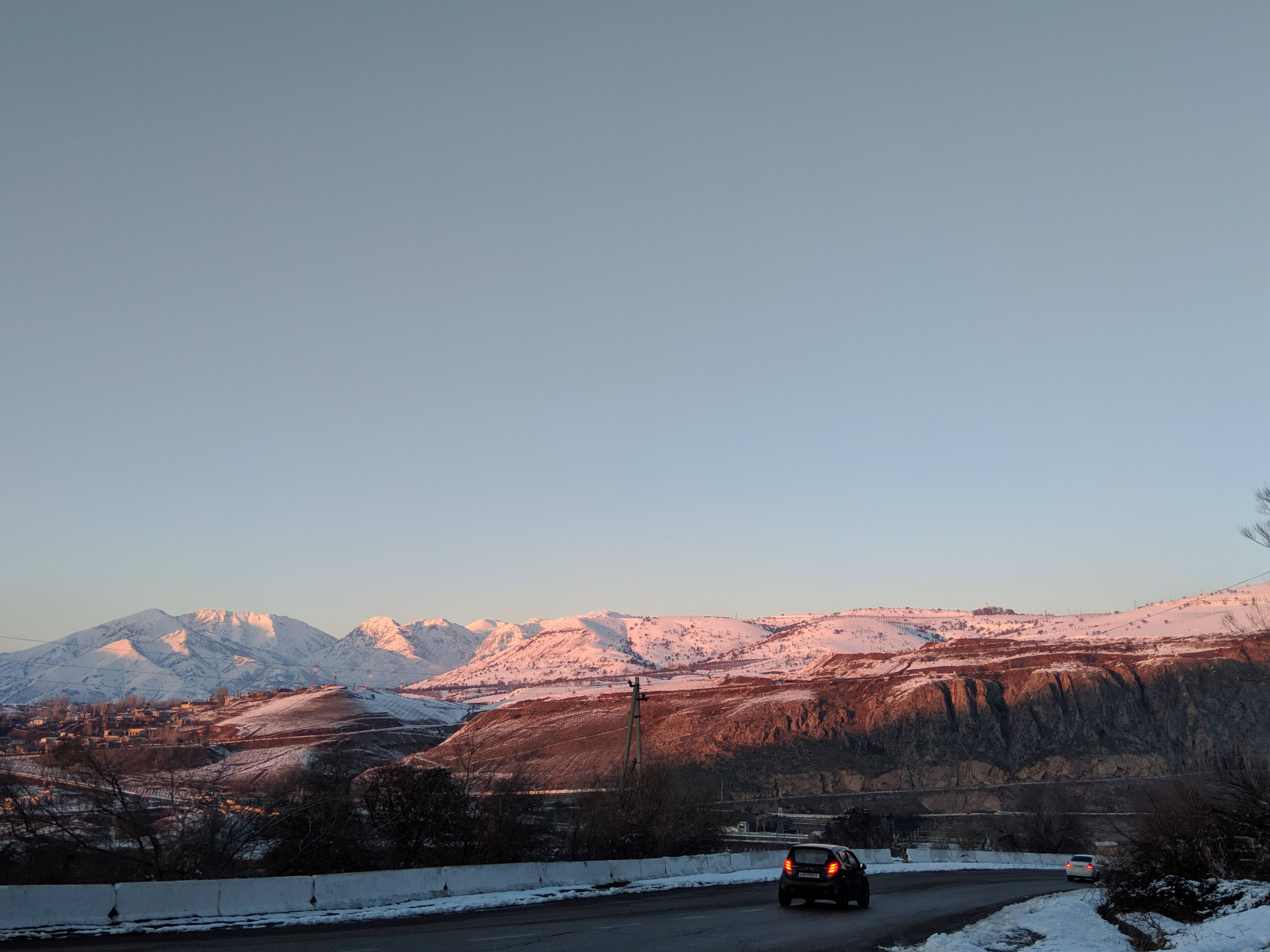 烏茲別克斯坦安集延雪山景觀 壁紙 — 一輛汽車沿著積雪的道路行駛，粉紅色和橙色的日落光芒以溫暖的色調沐浴著白雪覆蓋的山脈。