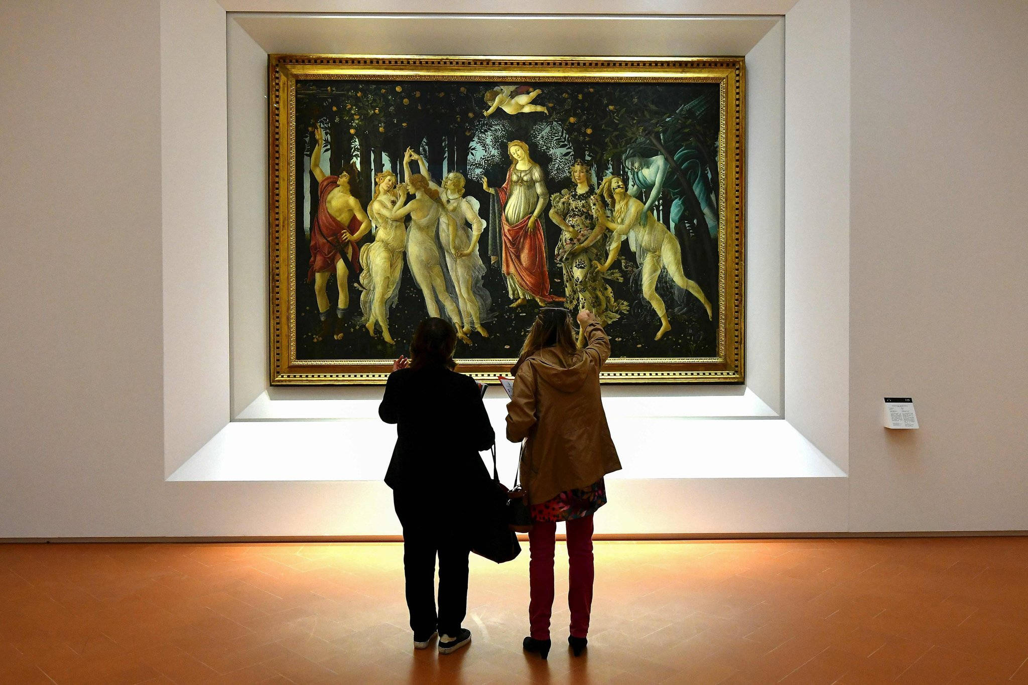 烏菲茲美術館波提切利《春》畫作 壁紙 — 兩位訪客在溫暖燈光的博物館畫廊中欣賞一幅大型華麗的文藝復興神話人物畫作。