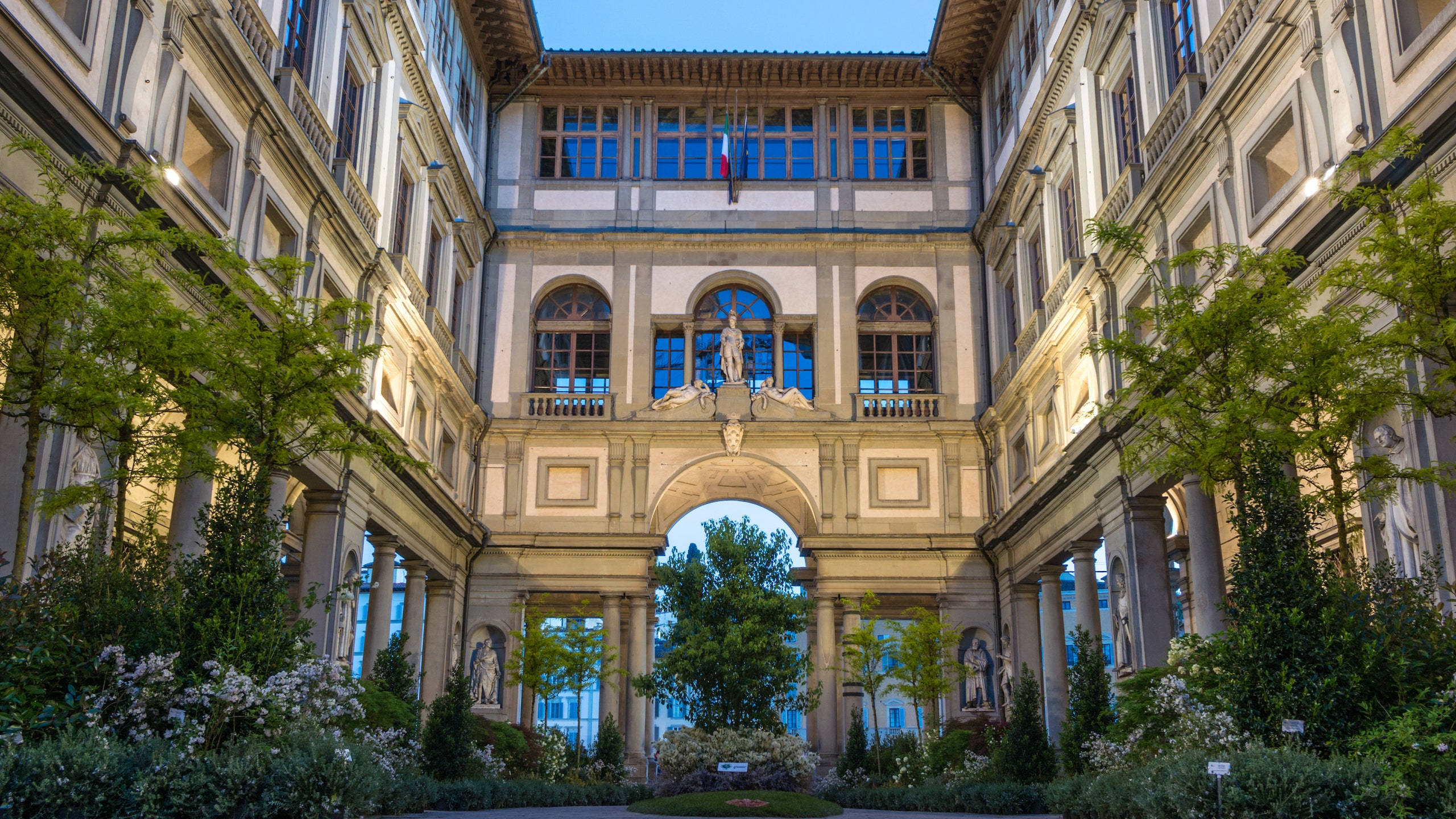 烏菲茲美術館與樹木 壁紙 — 一個宏偉的古典庭院，擁有對稱的建築、拱形走道和晴朗藍天下的茂密綠植。
