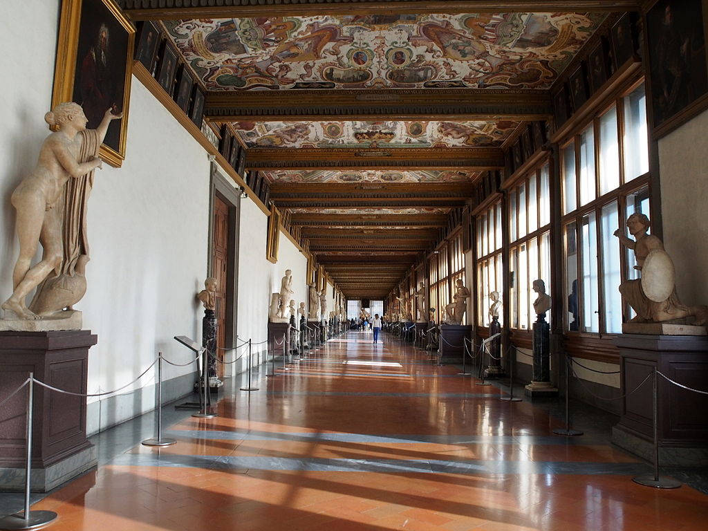烏菲茲美術館走廊 壁紙 — 一個宏偉的博物館畫廊，古典雕像排列在拋光地板上，上方是彩繪天花板，沐浴在自然光中。