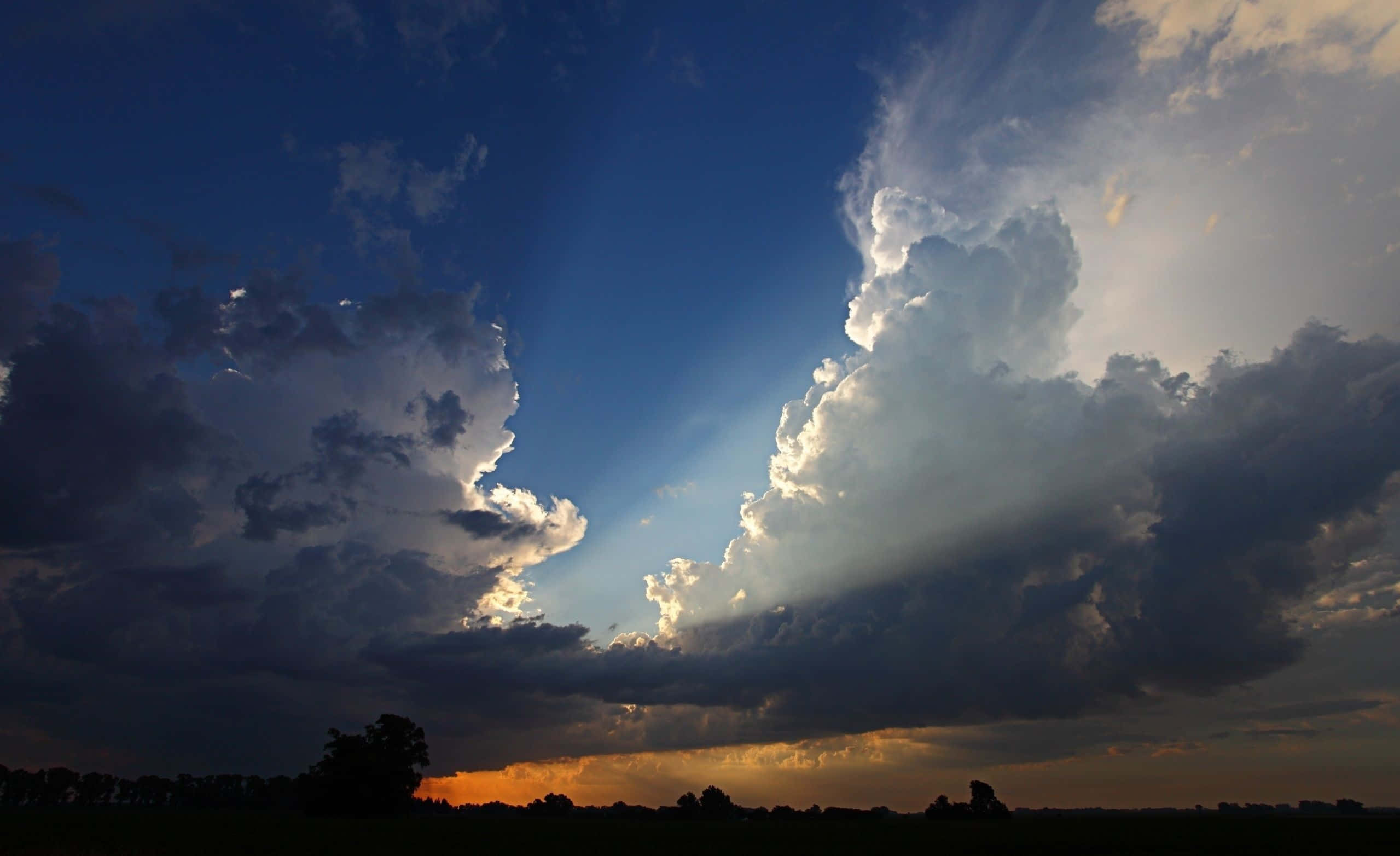 烏雲與陽光穿透的天體背景 壁紙 — 戲劇性的陽光穿透高聳的風暴雲層，照射在日落時分剪影般的田野上。