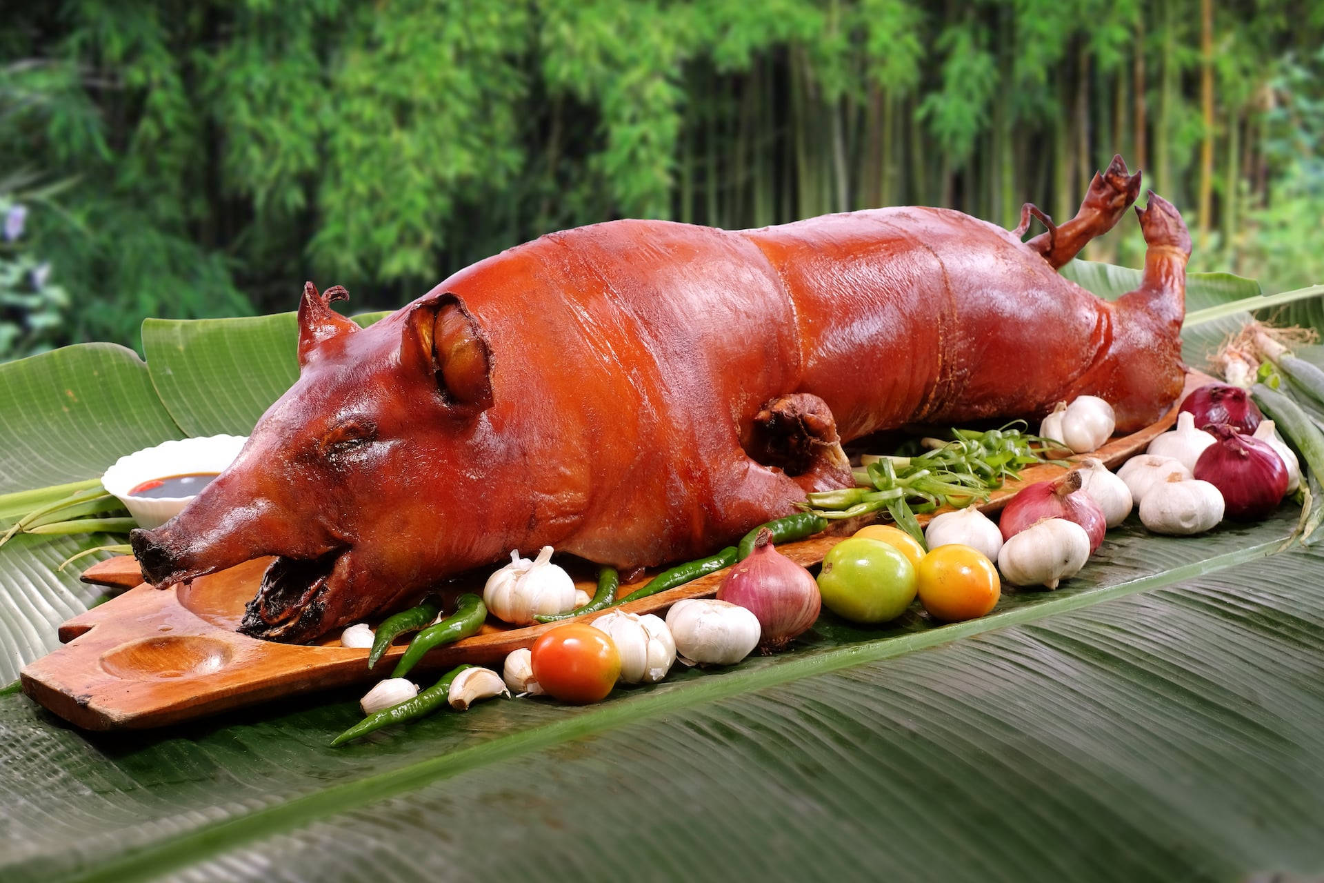烤乳豬包餐 壁紙 — 一隻烤全豬，金黃酥脆的豬皮，放在香蕉葉上，周圍有蒜頭、辣椒和番茄。