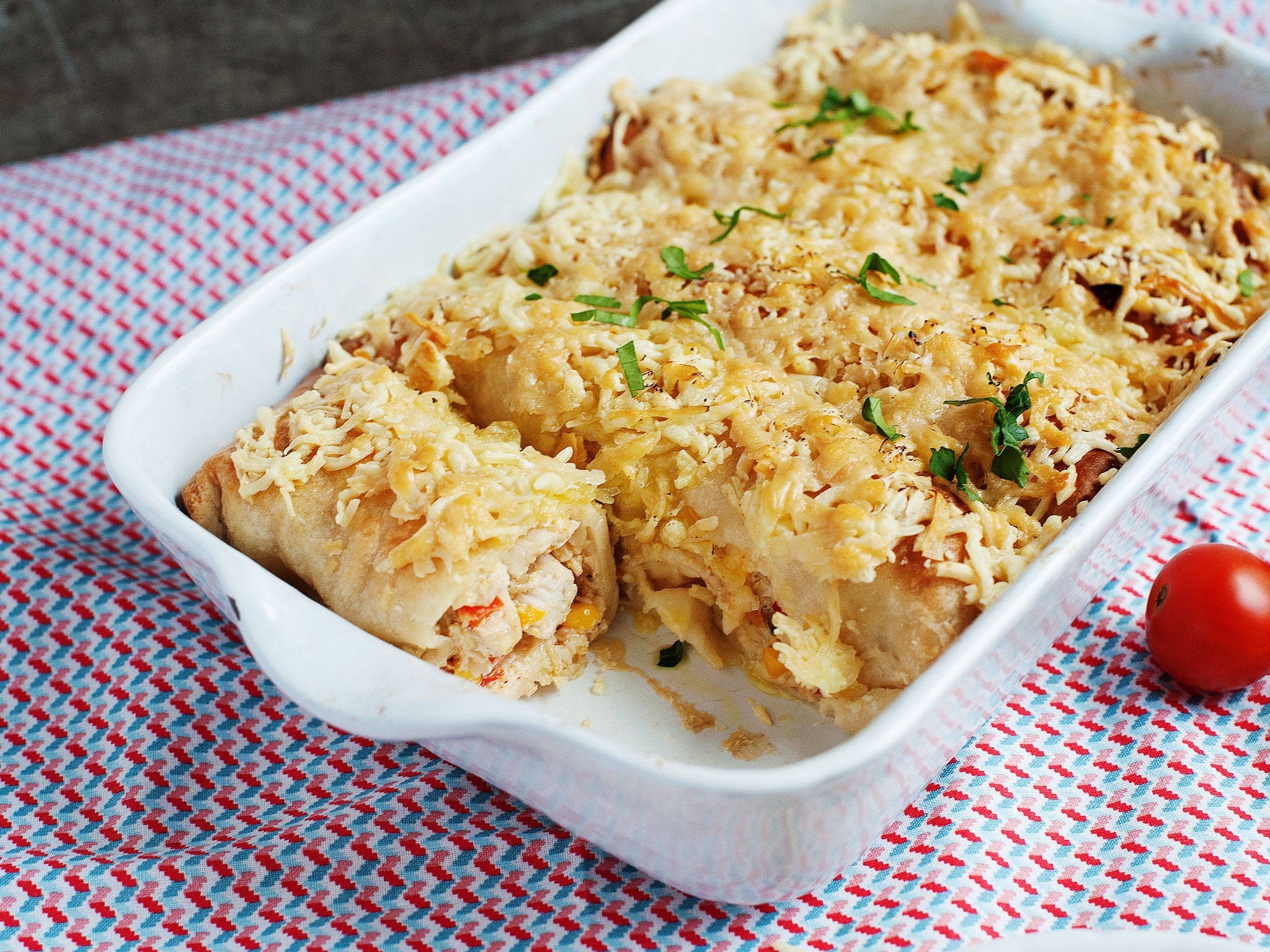 烤奶酪 enchiladas 盤 壁紙 — 金黃色芝士雞肉玉米捲餅放在白色烤盤中，置於紅藍格子布上，旁邊有一顆櫻桃番茄。