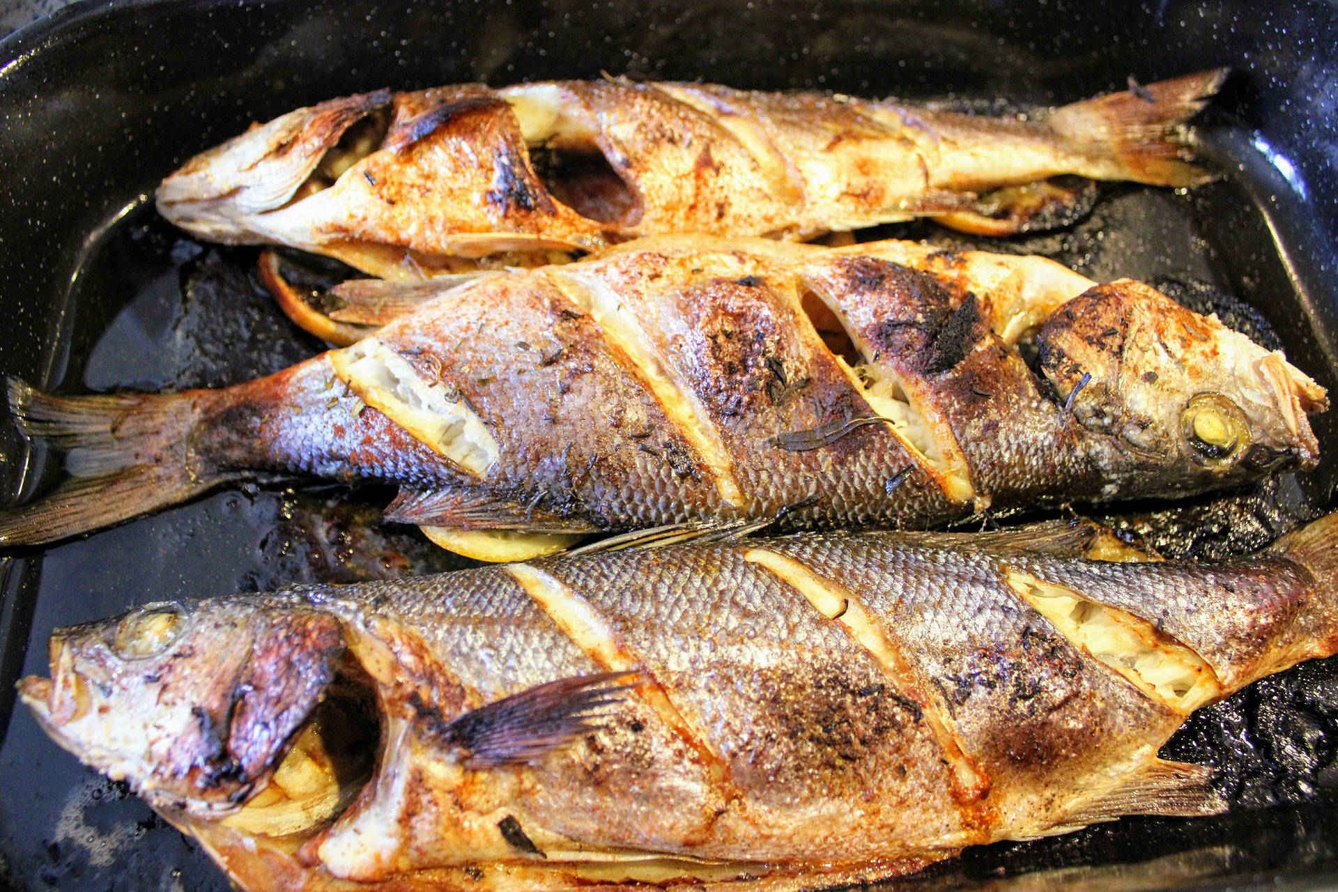 烤海鰱魚 壁紙 — 三條烤全魚，金黃色的魚皮和檸檬片在深色平底鍋中滋滋作響。