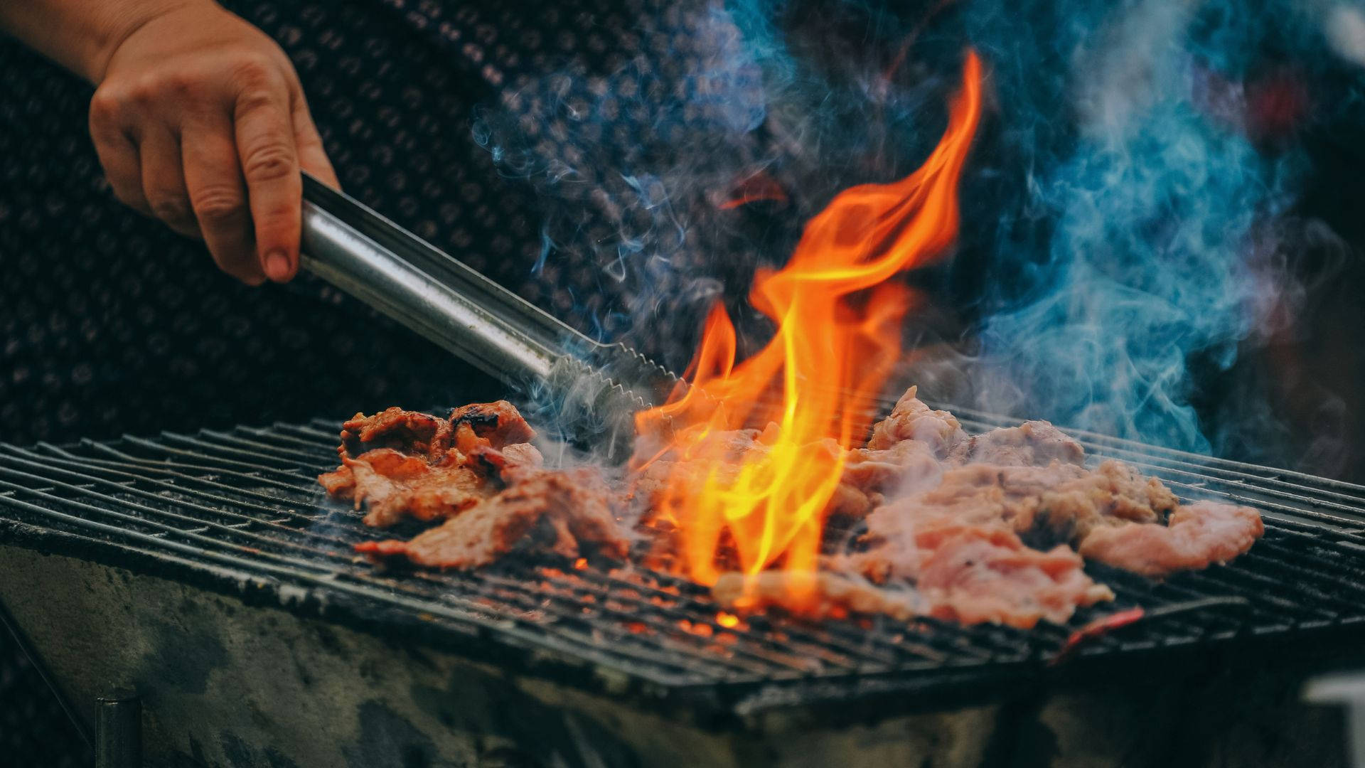 烤肉熱力 壁紙 — 一隻手用鉗子將在炭火烤架上滋滋作響的肉翻面，下方是鮮豔的橙色火焰，煙霧裊裊升起。