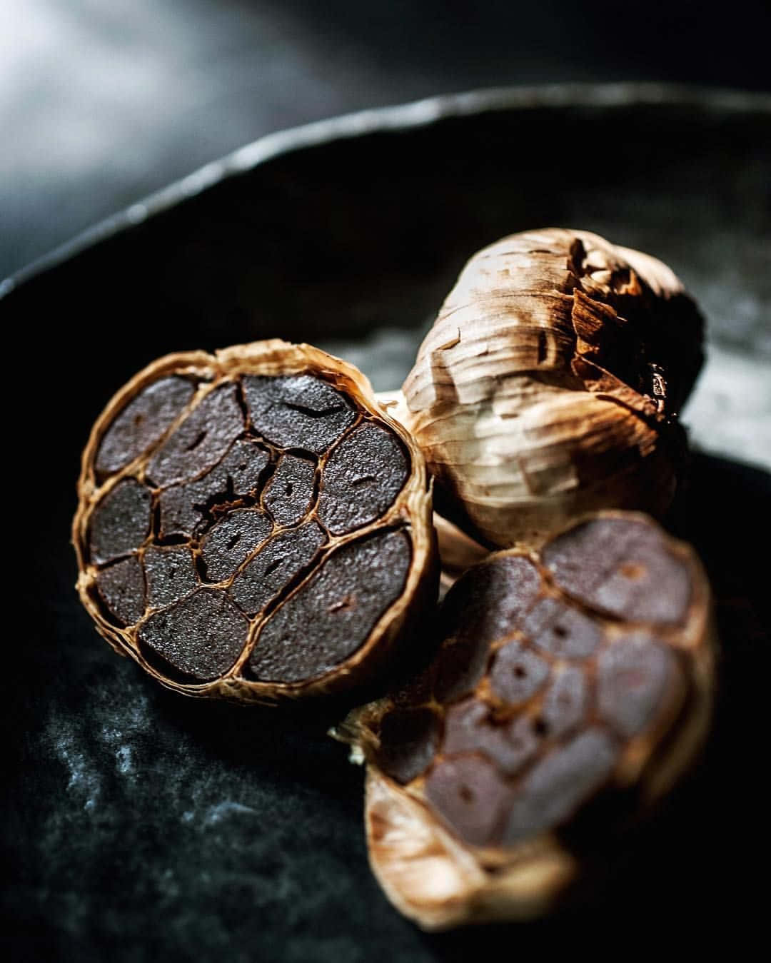 Black Garlic 桌布 壁紙 — 烤蒜瓣的特寫，內部焦黑，放在深色啞光碗中。