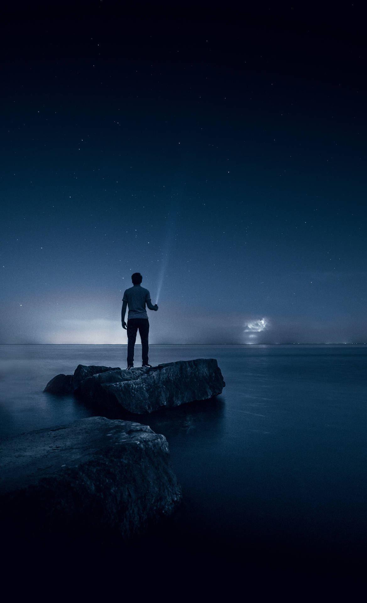夜晚站在岩石上的人將手電筒照向水面上方的星空