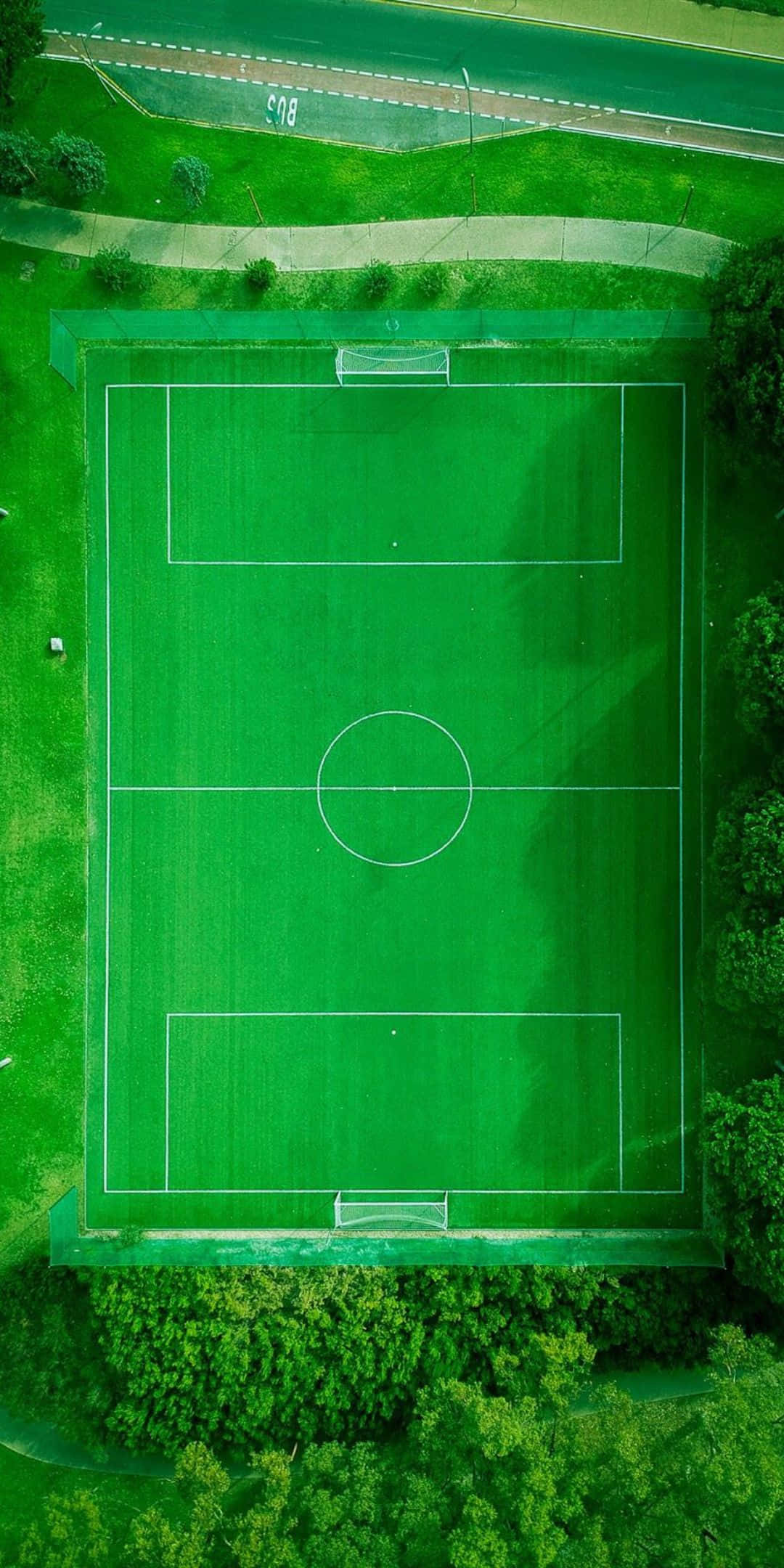 無人機拍攝足球場 Pixel 3 足球背景 壁紙 — 鳥瞰圖：一片生機勃勃的綠色足球場，四周環繞著茂密的樹木，並毗鄰一條道路。