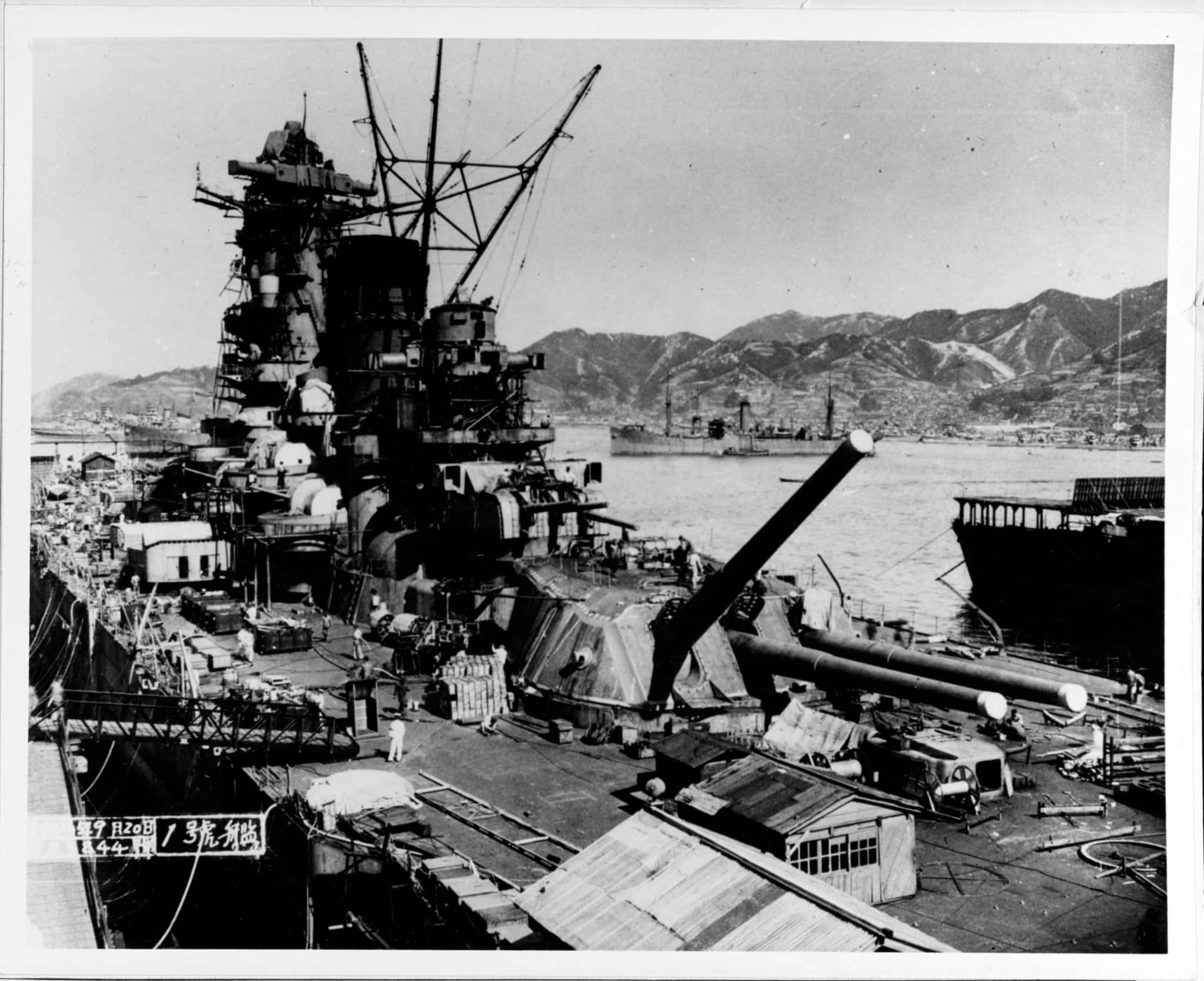 無垠海域的壯闊戰場！ 壁紙 — 一艘巨大的戰艦主宰著港口，背景有山脈，甲板上可見船員。