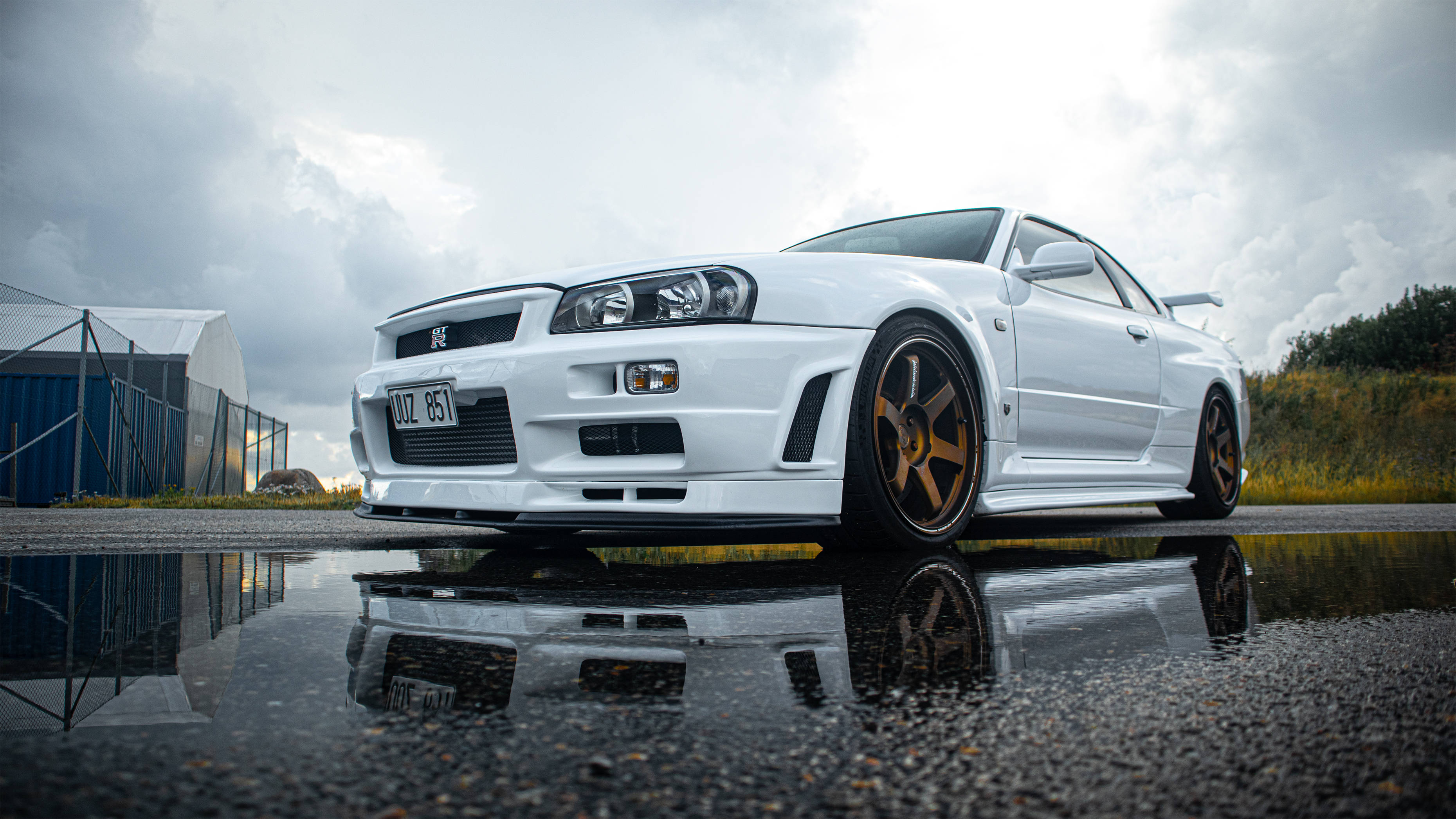 無敵珍珠白Nissan Skyline GTR R34 壁紙 — 一輛白色日產Skyline R34跑車配金色輪圈，在陰天下倒映在水窪中。