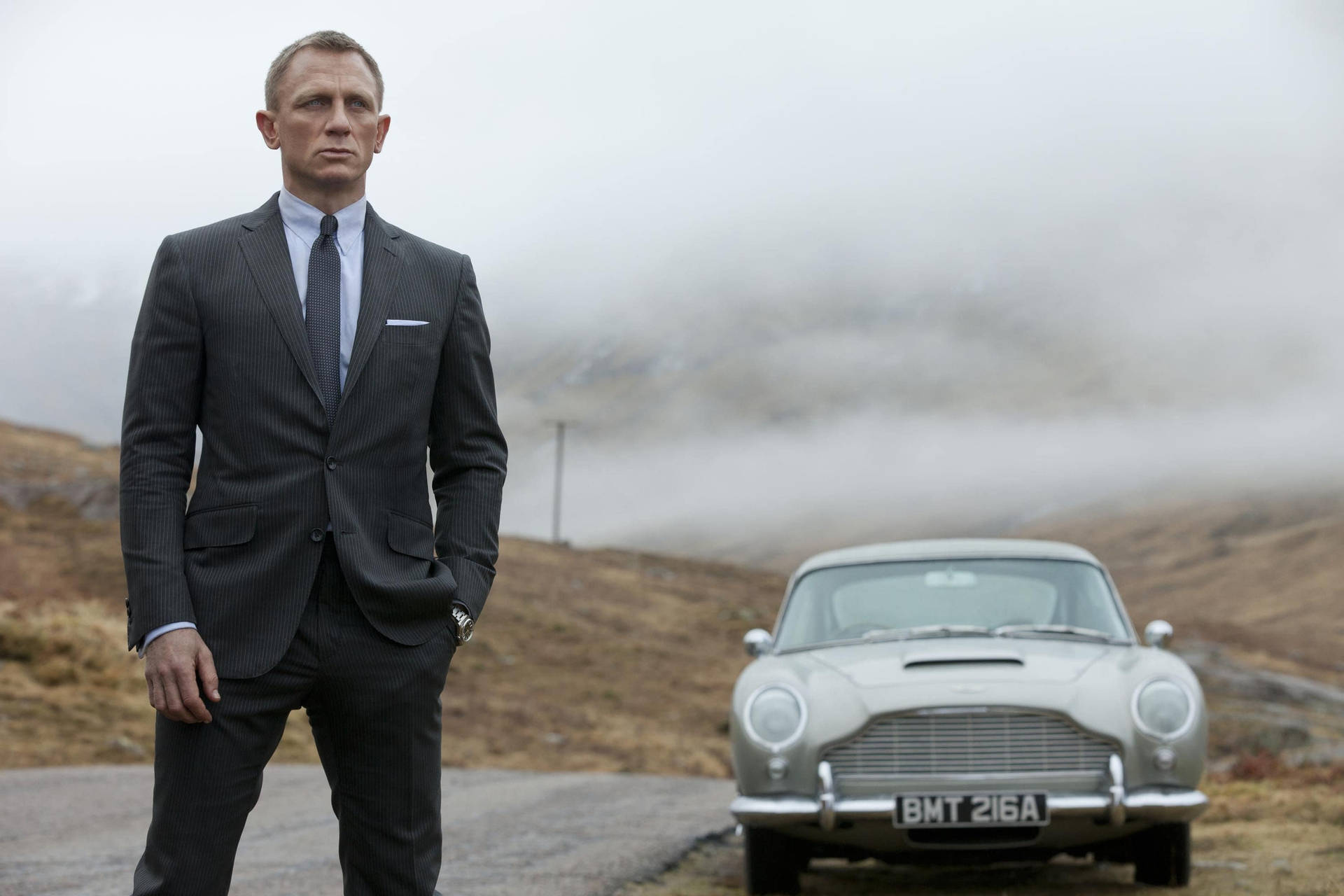 身穿西裝的男子在霧氣瀰漫的山路上站在銀色Aston Martin旁