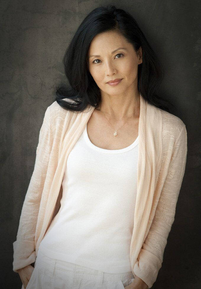 無瑕之美：Tamlyn Tomita 的肖像 壁紙 — 一位優雅的黑髮女性，在紋理灰色背景前，穿著柔軟的桃色開襟衫，內搭白色上衣。
