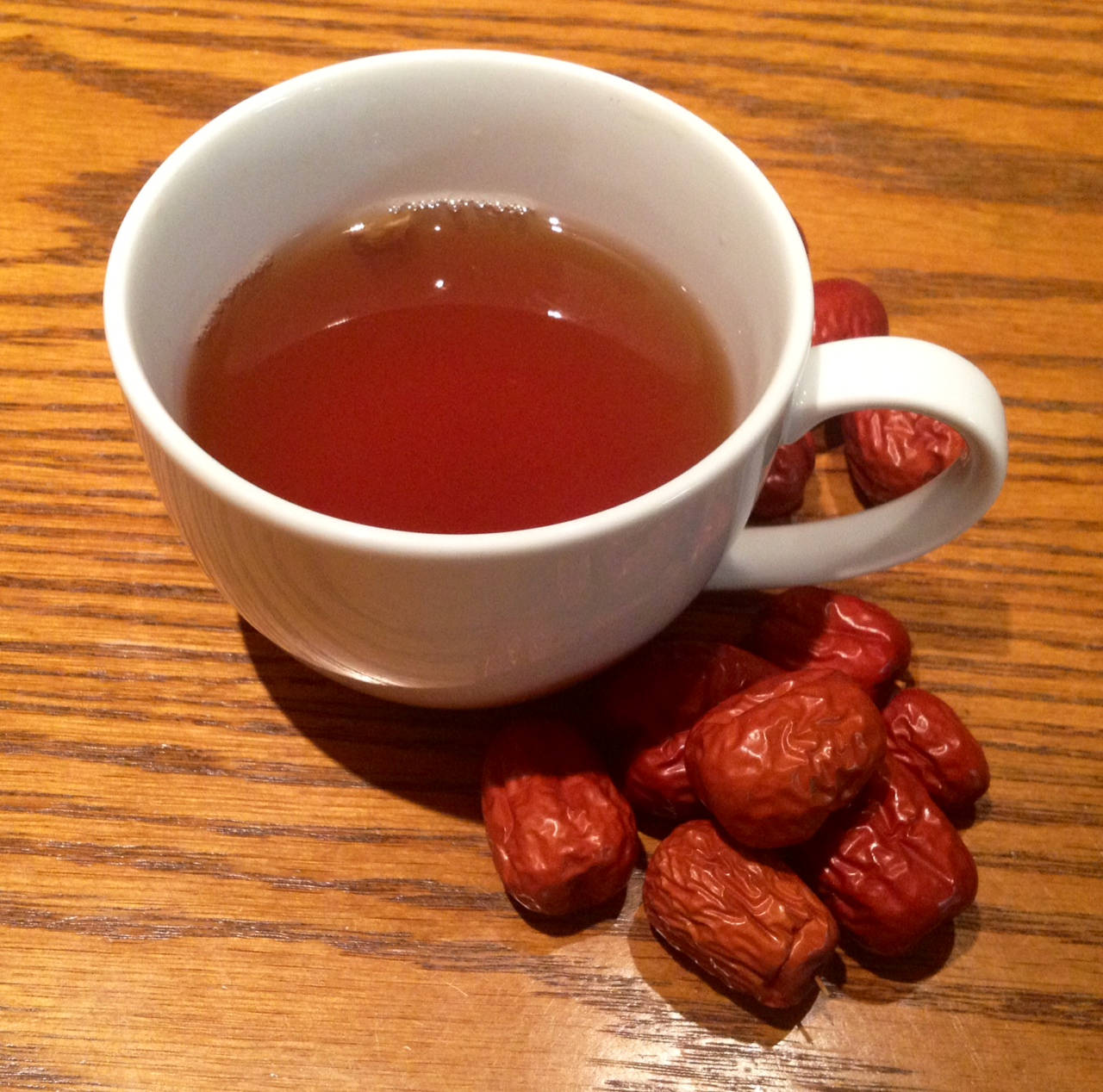 無花果種子與茶杯 壁紙 — 一個白色陶瓷杯裝滿琥珀色茶，放在溫暖的木桌上，旁邊是飽滿的紅棗。