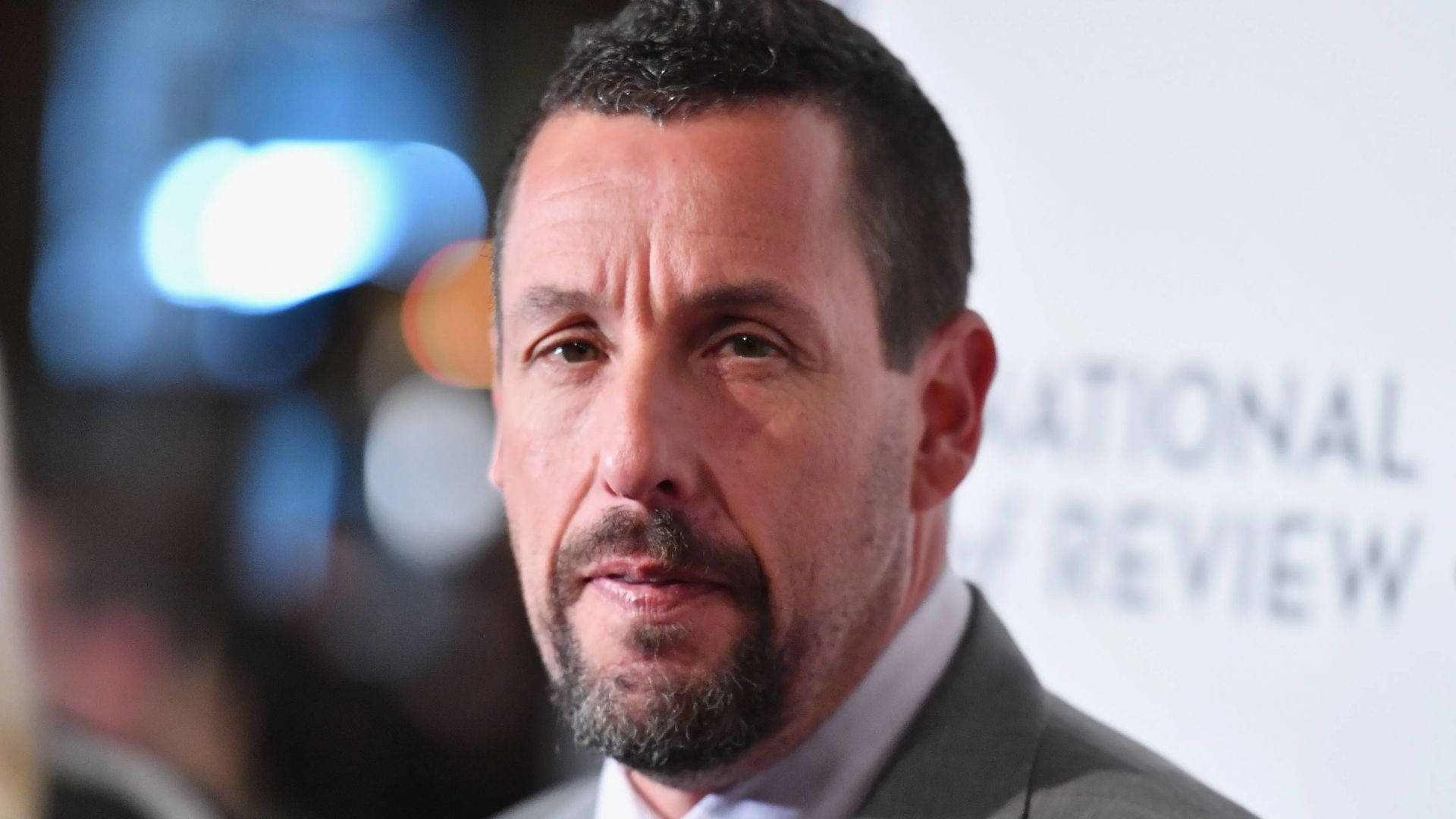 無表情亞當·桑德勒 壁紙 — Adam Sandler 喺正式活動嘅特寫肖像，背景有模糊燈光同 'NATIONAL REVIEW'。