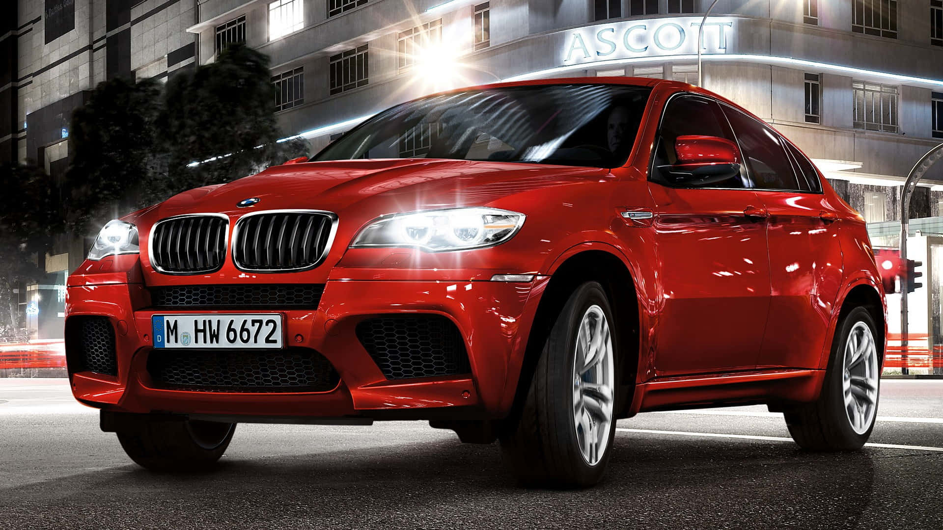 無論追求動力或奢華，BMW X6 M 全部兼備 壁紙 — 一輛光澤的紅色BMW X6運動型多功能車停在夜晚潮濕的城市街道上，周圍有燈火通明的建築物和發光的車頭燈。