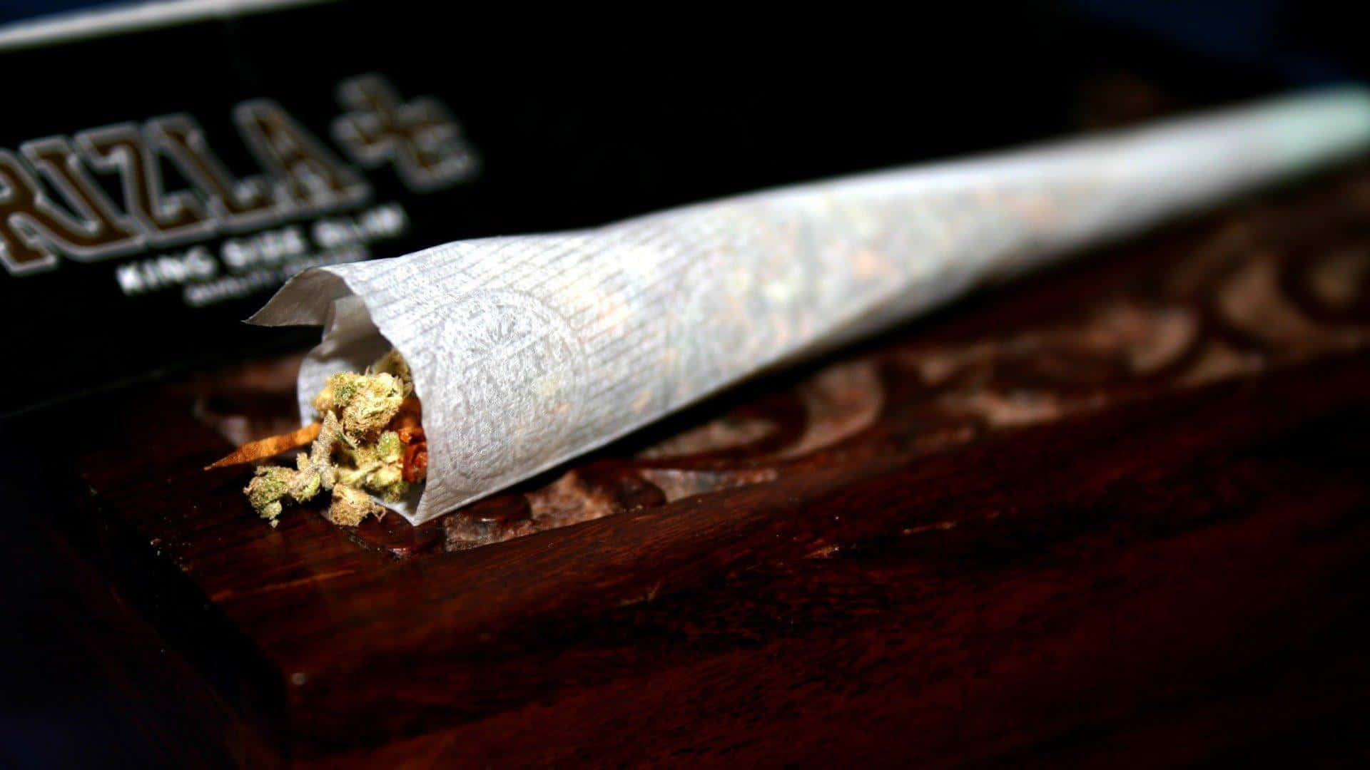 煙卷末端露出大麻 壁紙 — 一個捲好的大麻煙捲特寫，放在深色木質表面上，背景有一個品牌盒子。