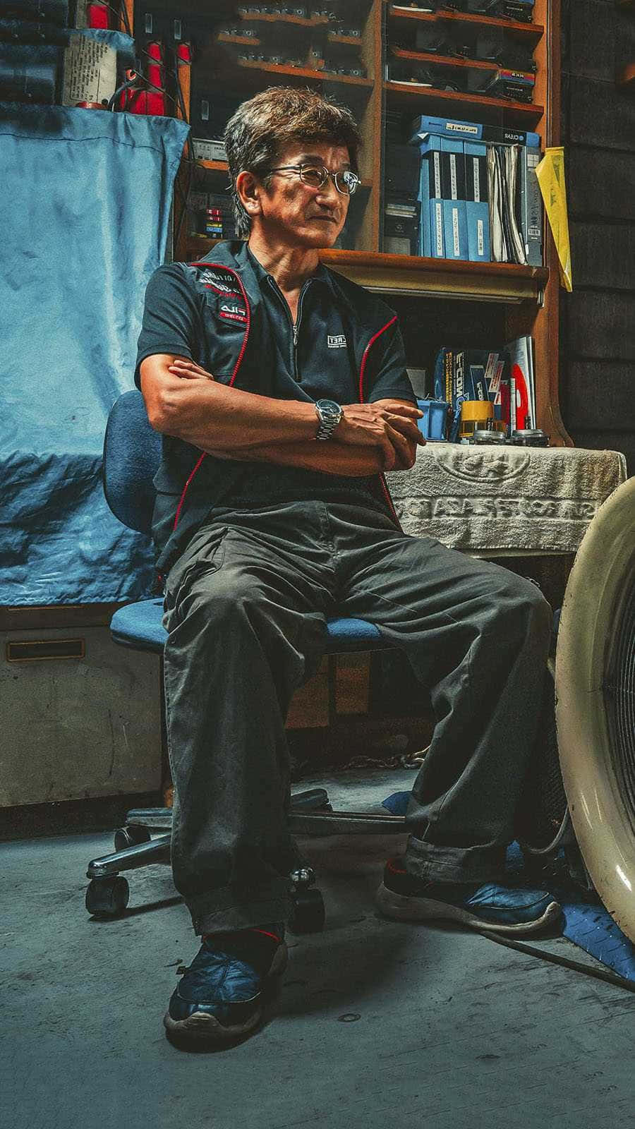 煙霧 Nagata 坐在修理店內 壁紙 — 一位年長男子穿著賽車制服，自信地坐在雜亂的車庫中，身後有工具和架子。