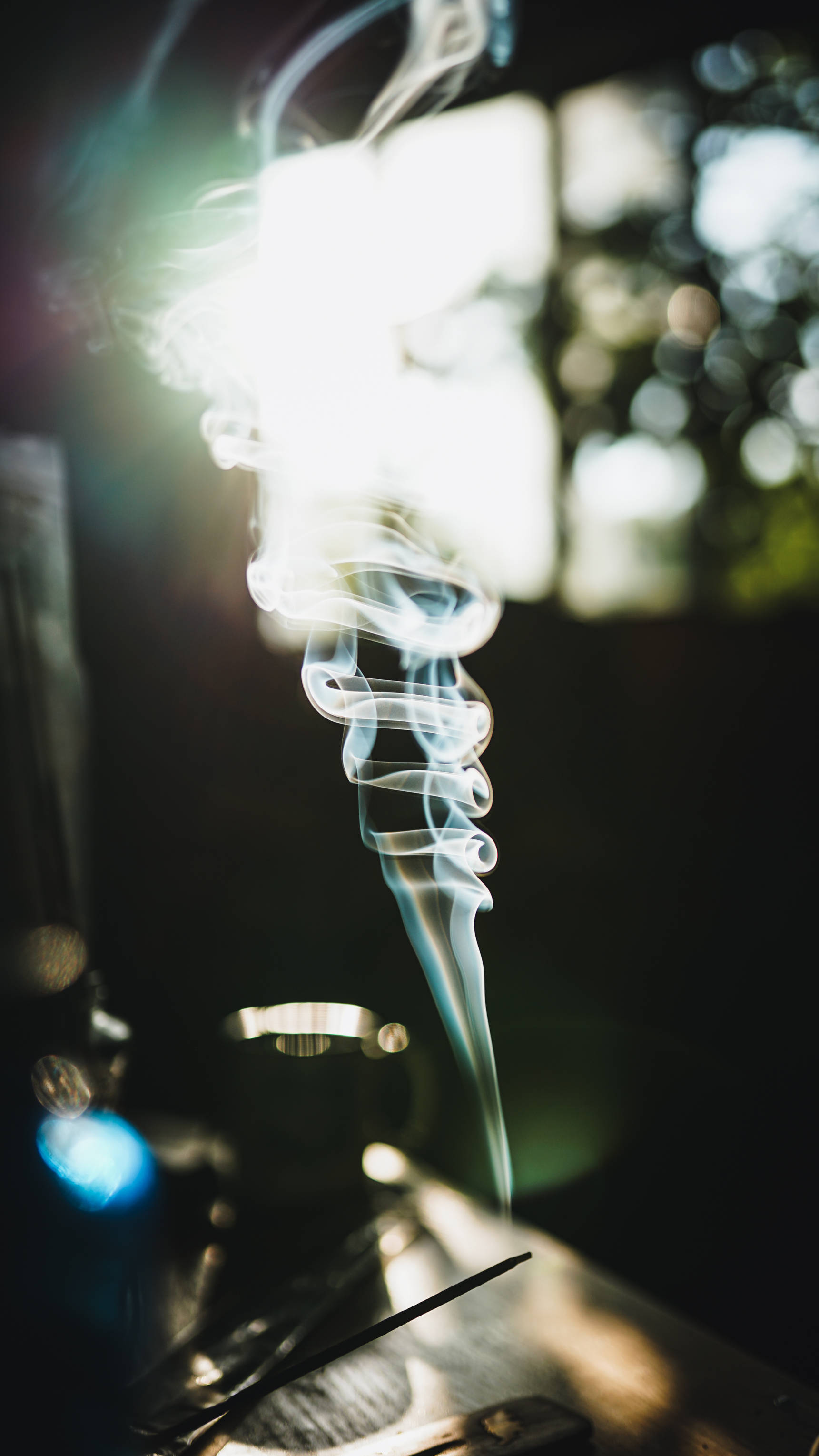 煙霧攝影 HD 壁紙 — 一縷纖細的白煙螺旋從黑暗表面升起，背景模糊帶有柔和散景光點。