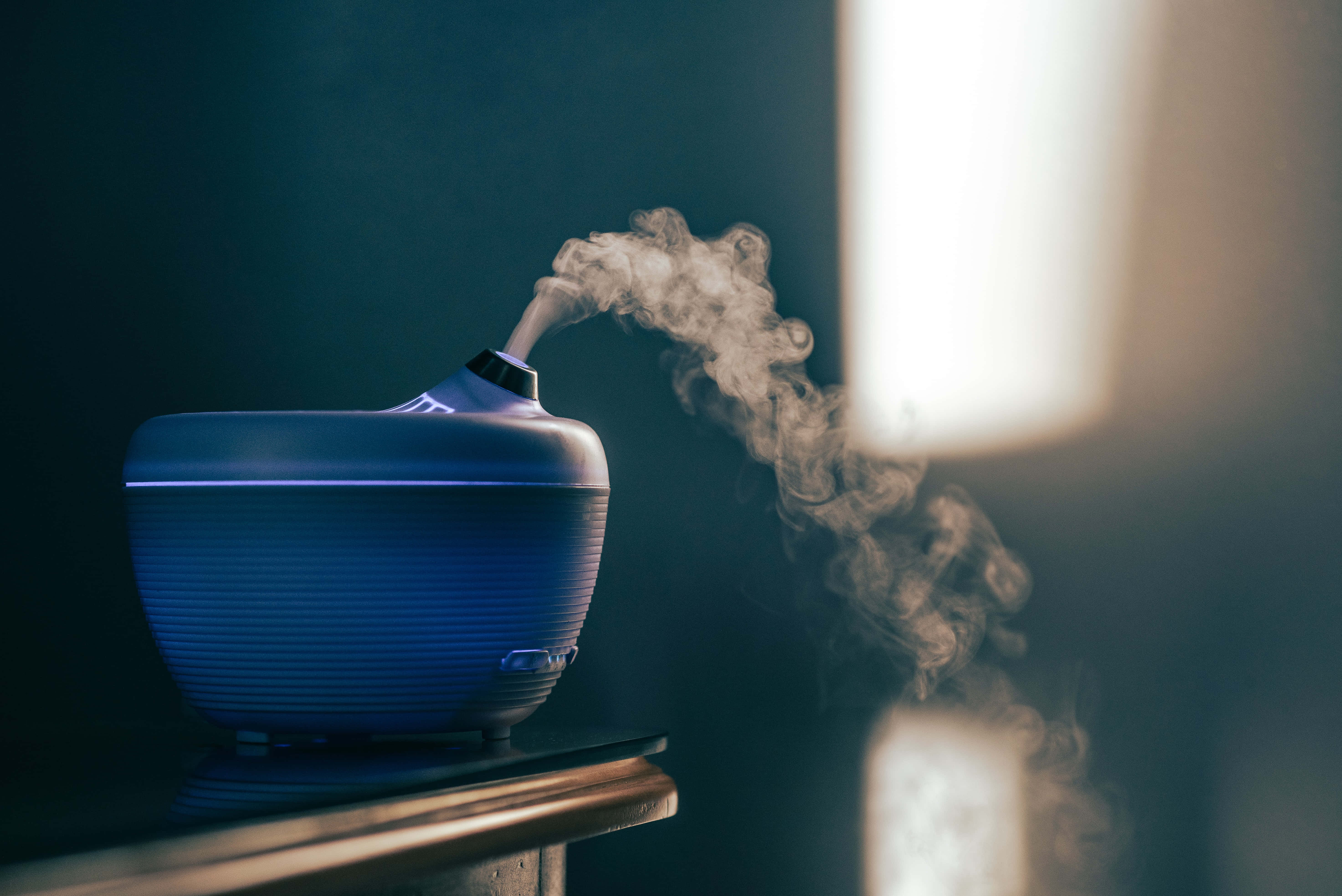 煙霧灰調的抽象風景 壁紙 — 一部時尚的藍色加濕器在昏暗的房間中釋放出溫和的霧氣，柔和自然光透過窗戶照射進來。