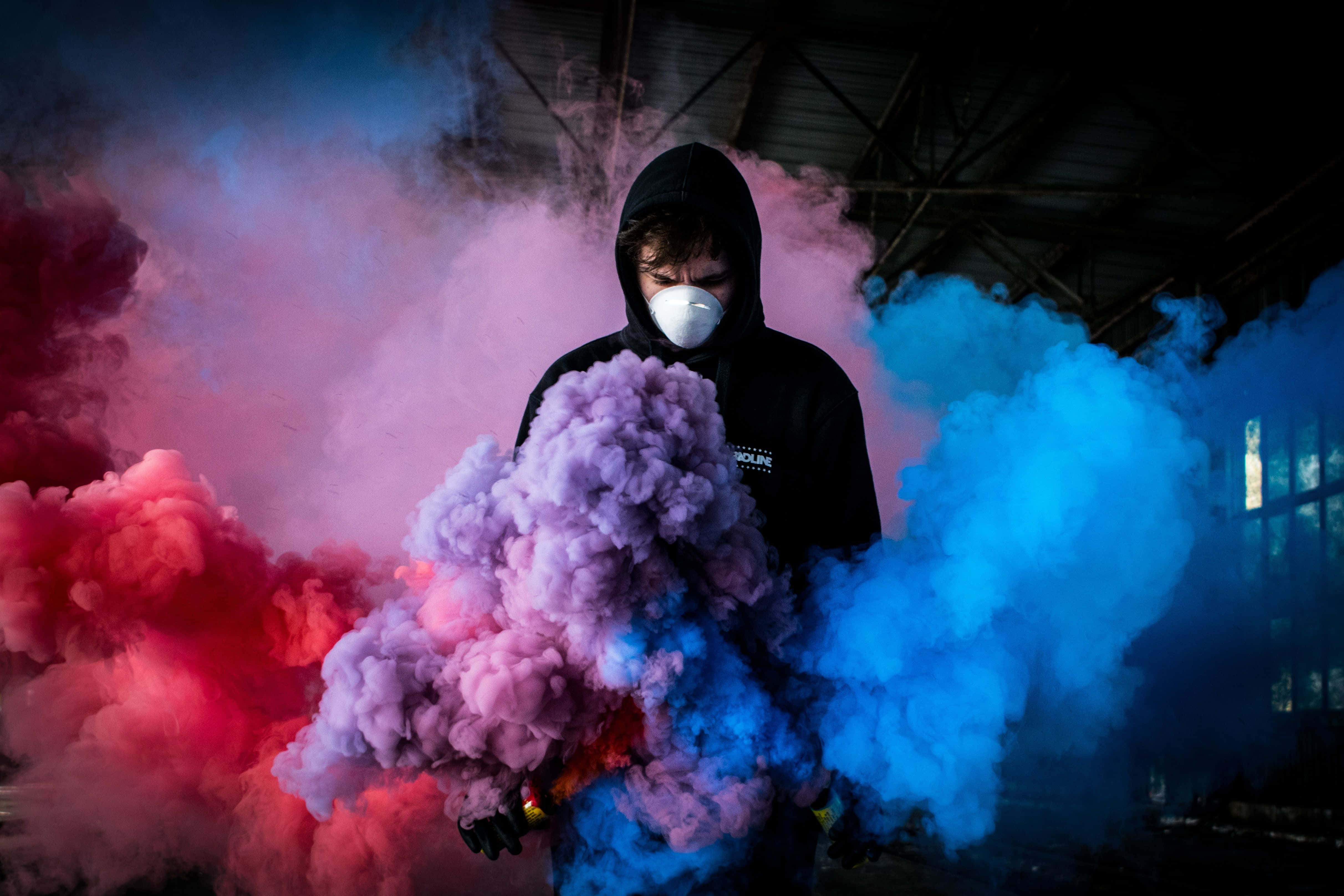 煙霧炸彈照片中的男子 壁紙 — 一個穿著連帽衫和口罩的人站在昏暗的工業空間中，周圍環繞著旋轉的紅色、紫色和藍色煙霧。