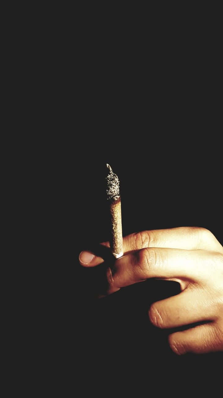煙頭的煙草捲煙 壁紙 — 一隻手拿著點燃的香煙，背景為純黑色，戲劇性的燈光突出了煙霧和手指。