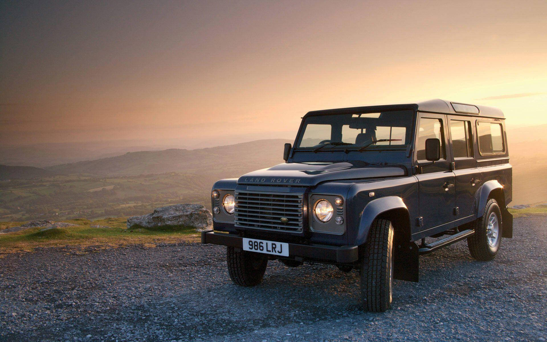 深藍色 Land Rover Defender 停泊在山丘上，日落時分，背景是連綿起伏的山丘。