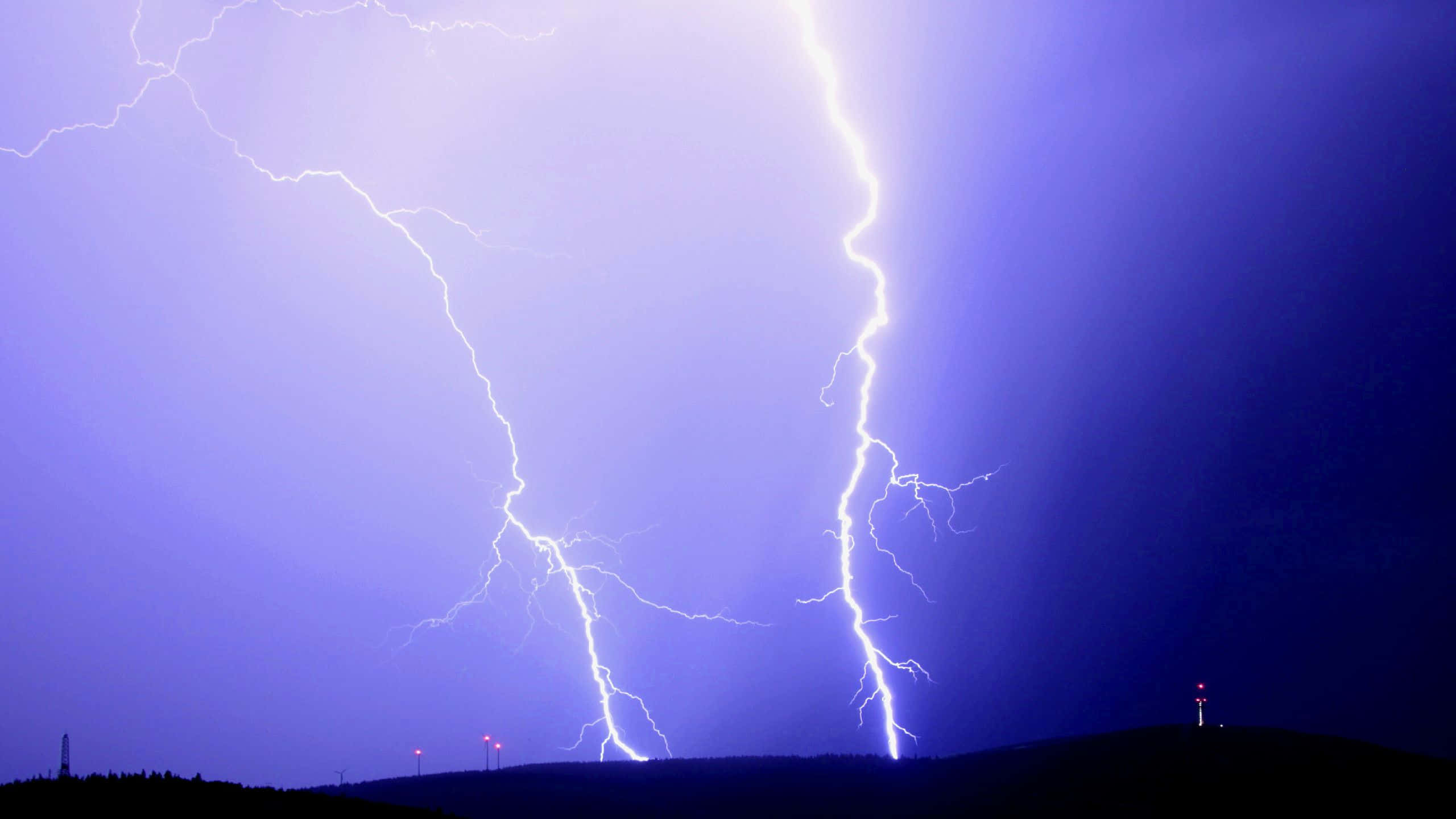 照亮夜空的閃電 壁紙 — 兩道巨大的閃電劃破深紫色暴風雨的天空，下方是黑暗輪廓的山丘，遠處有紅燈。