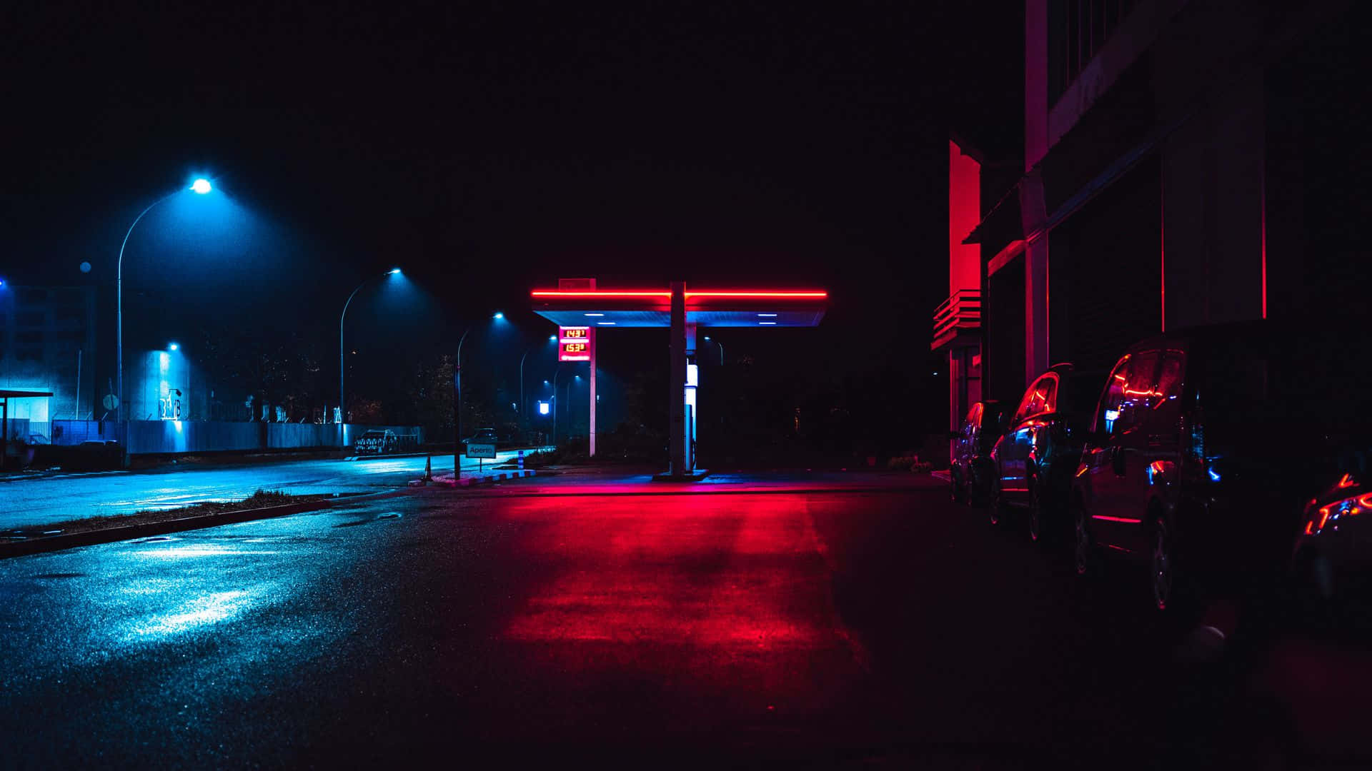 照亮的夜之城 壁紙 — 夜晚霓虹燈照亮的加油站，紅色和藍色的燈光反射在潮濕的路面和停放的汽車上。