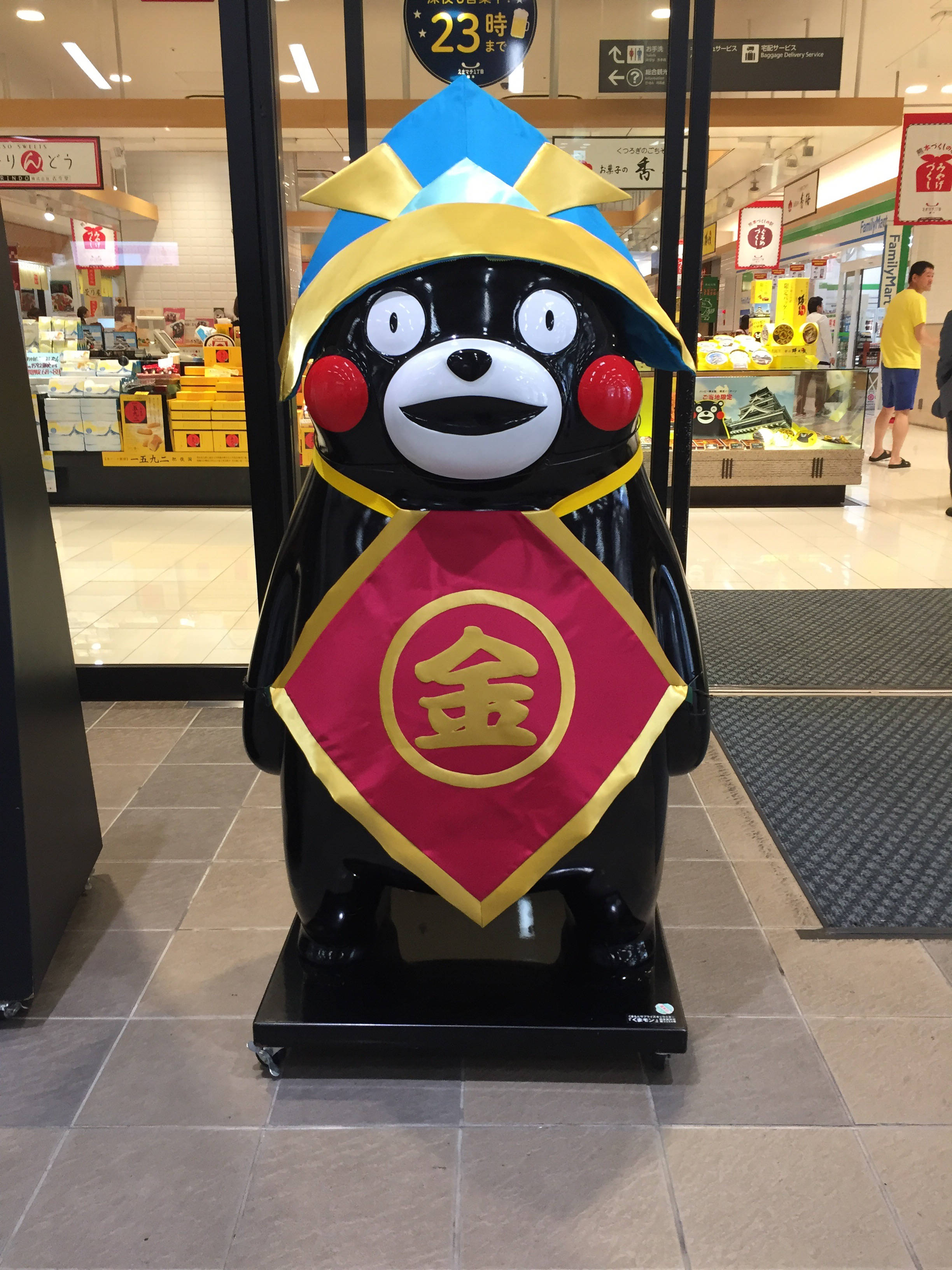 熊本熊商店立牌 壁紙 — 一尊光滑的黑色熊本熊雕像，戴著藍黃色帽子和帶有漢字的紅金色腰帶，站在光線明亮的商店通道中。