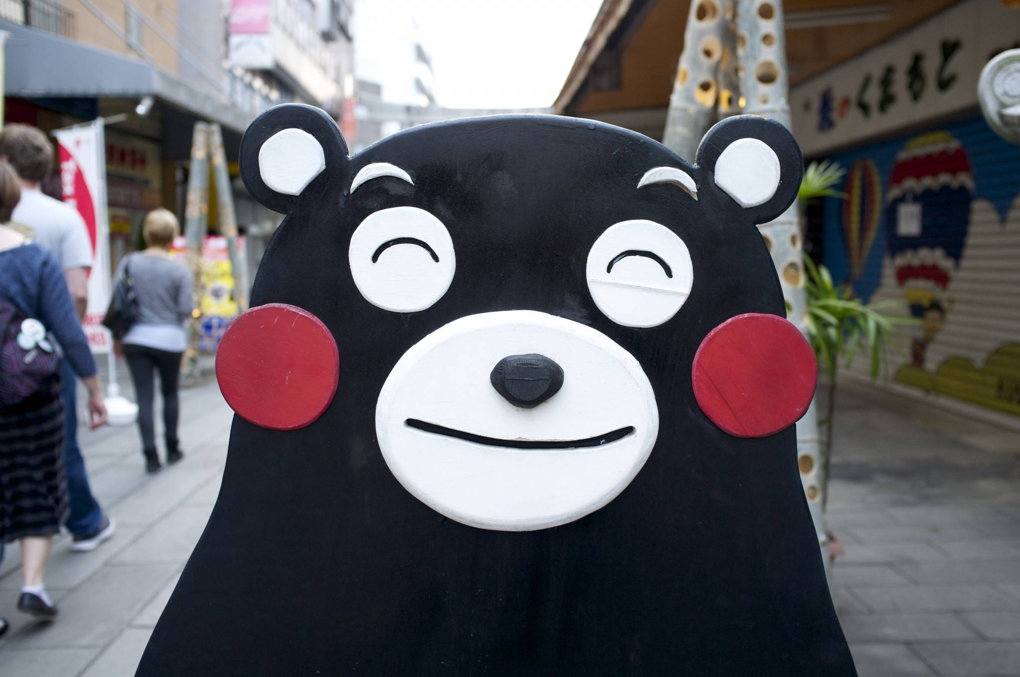 熊本熊戶外立牌 壁紙 — 一隻大型黑色熊吉祥物，臉頰紅潤、雙眼緊閉，站在繁忙的城市人行道上，行人來來往往。
