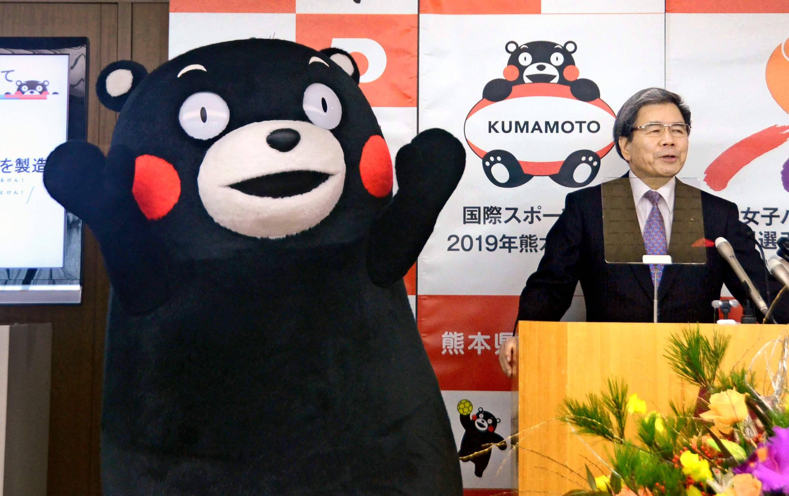 熊本熊與熊本縣知事 壁紙 — 一名穿西裝的男子在活動中於講台旁發言，旁邊有一個大型 Kumamon 吉祥物在揮手。