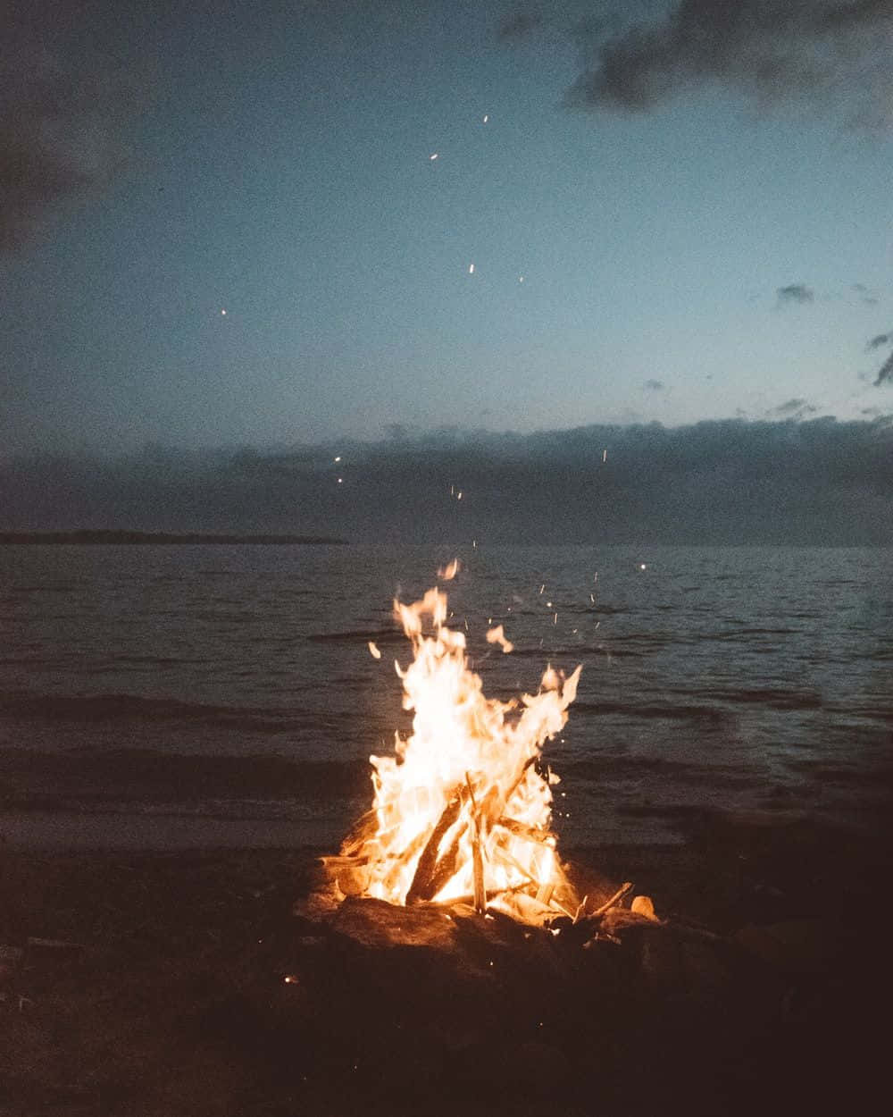 Beach Bonfire 桌布 壁紙 — 熊熊營火在海灘上暮色中閃爍，將溫暖的光芒投射在漸暗的海洋和多雲的天空上。