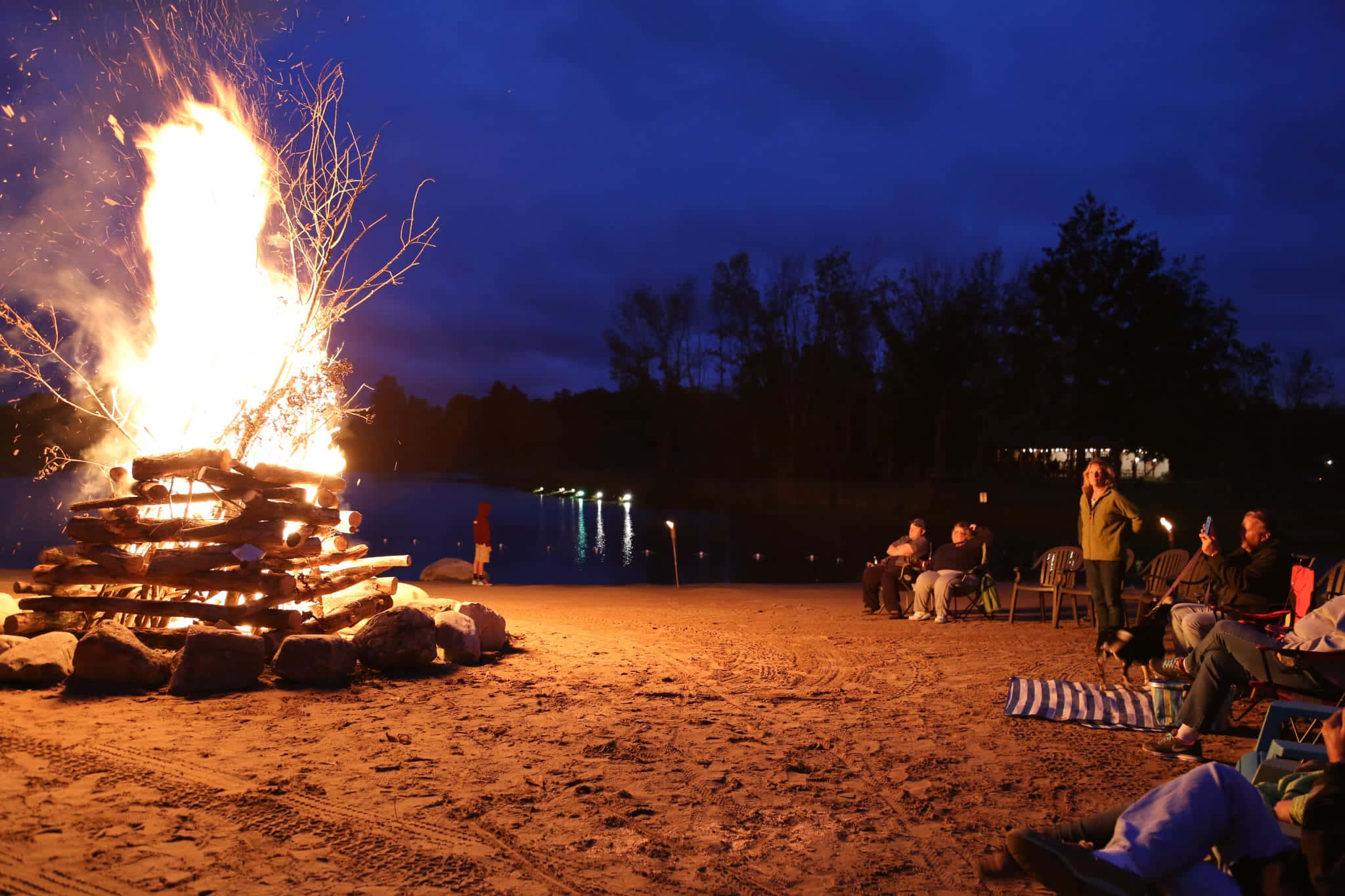 Beach Bonfire 桌布 壁紙 — 熊熊營火照亮了一群在暮色中於湖岸放鬆的人們，背景有樹木和遠處的燈光。