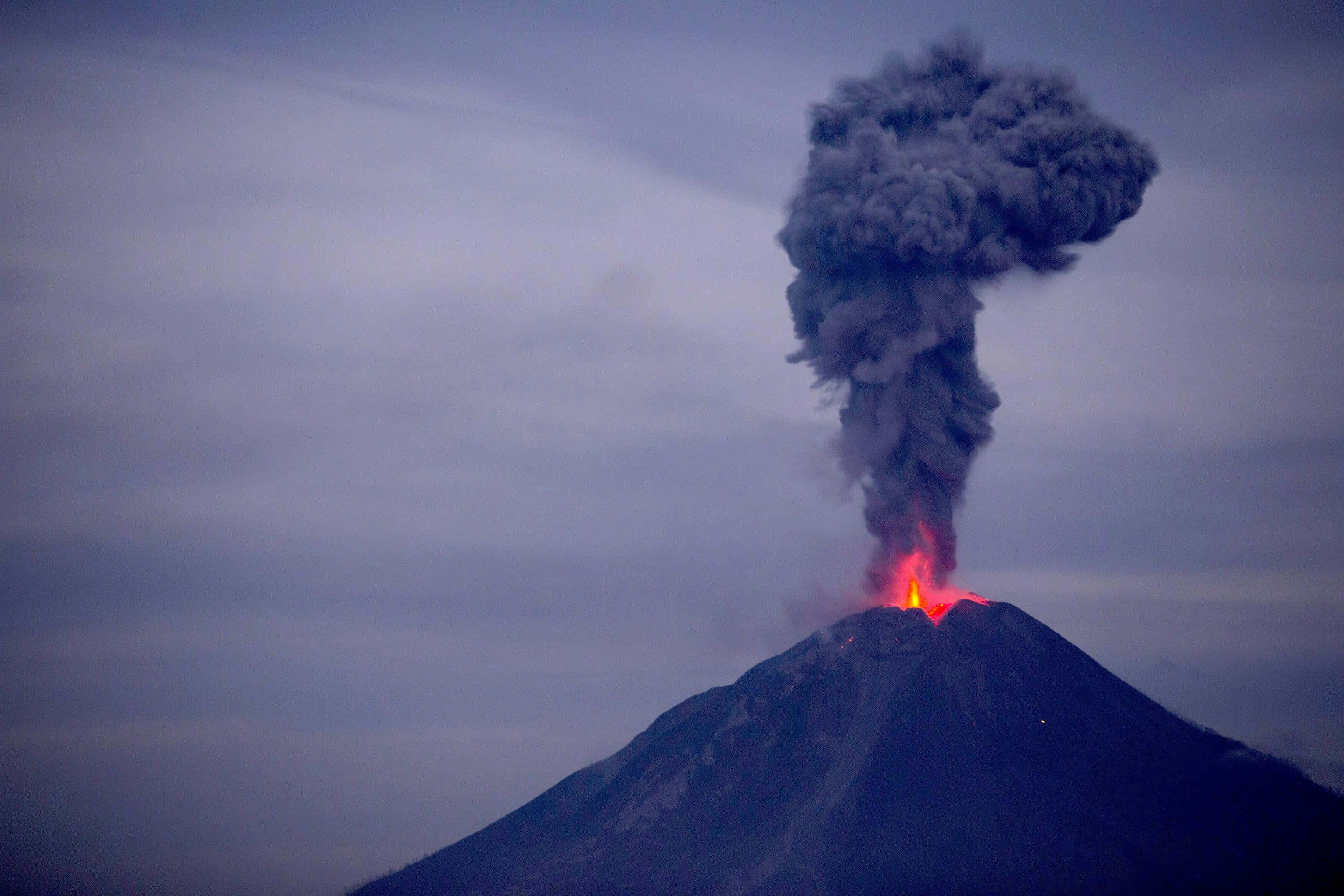 熔岩爆發，形態迷人，色彩繽紛 壁紙 — 一座火山爆發，噴出熾熱的熔岩和巨大的黑煙柱，映襯著暮色天空。