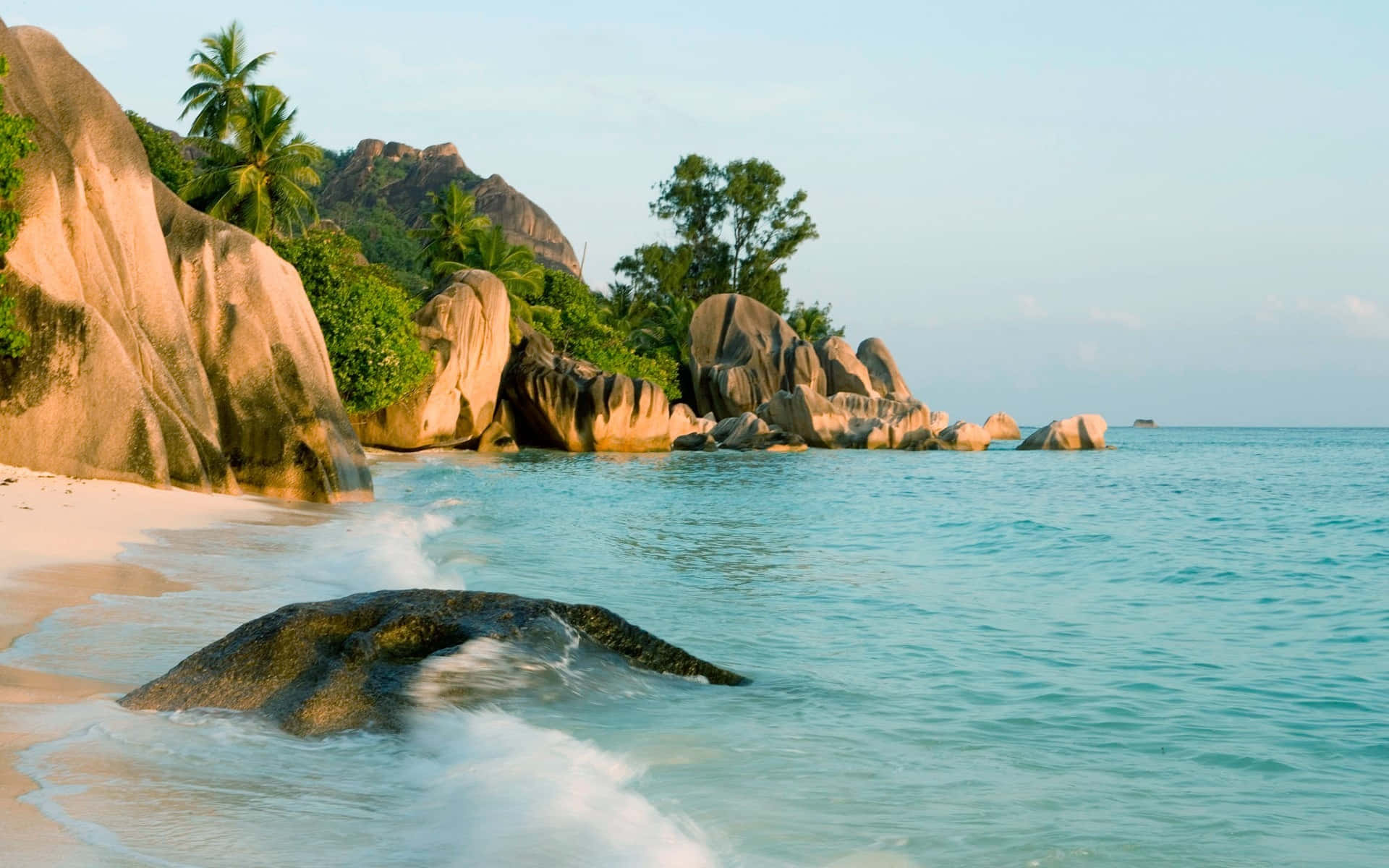 Seychelles Beach 桌布 壁紙 — 熱帶海灘，碧綠海水拍打巨岩，茂密棕櫚樹在晴朗天空下搖曳。