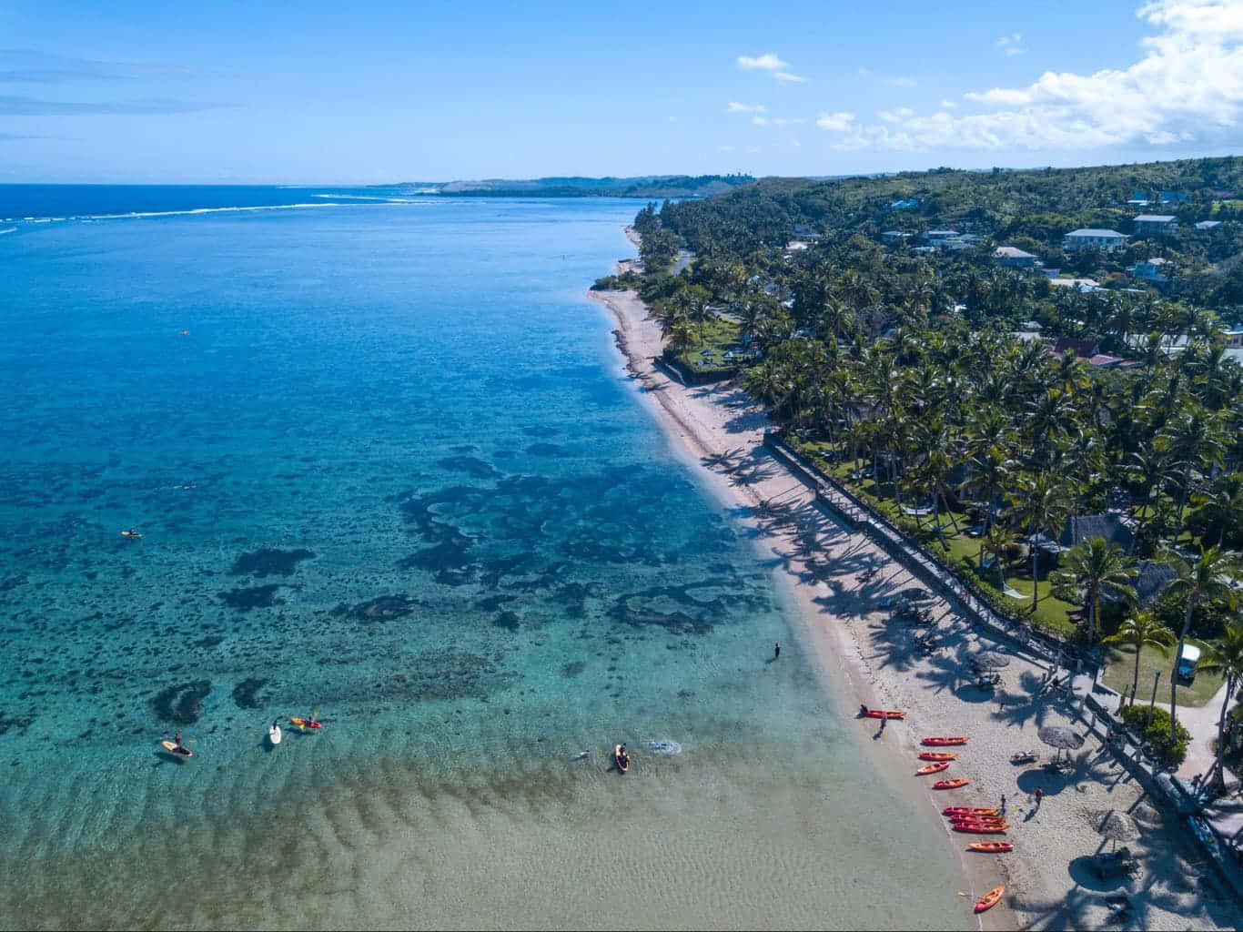 Fiji Beach 桌布 壁紙 — 熱帶海灘的鳥瞰圖，有碧綠的海水、珊瑚礁和棕櫚樹排列在海岸線上，還有皮划艇和人們享受陽光明媚的一天。