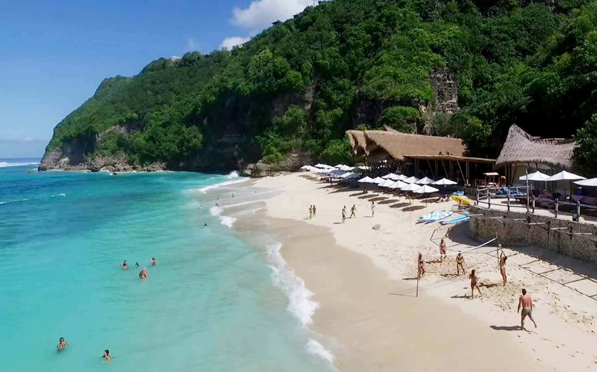 Bali 桌布 壁紙 — 熱帶海灘的鳥瞰圖，碧綠海水，人們在白色遮陽傘下游泳和打排球，旁邊是茂密的綠色懸崖。